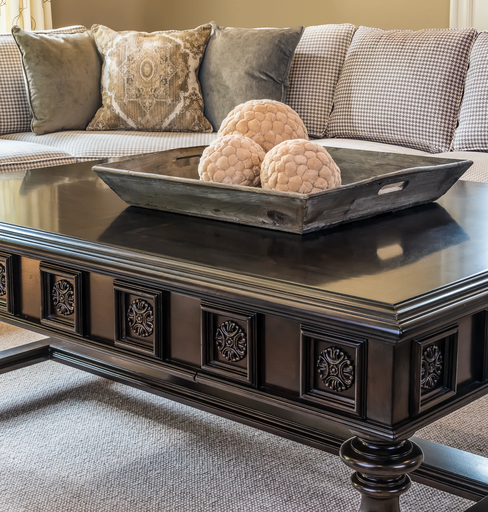 A dark, glossy wood coffee table with carved paneling is centered, holding a weathered wooden tray and three decorative spheres. A sofa with patterned pillows is in the background.