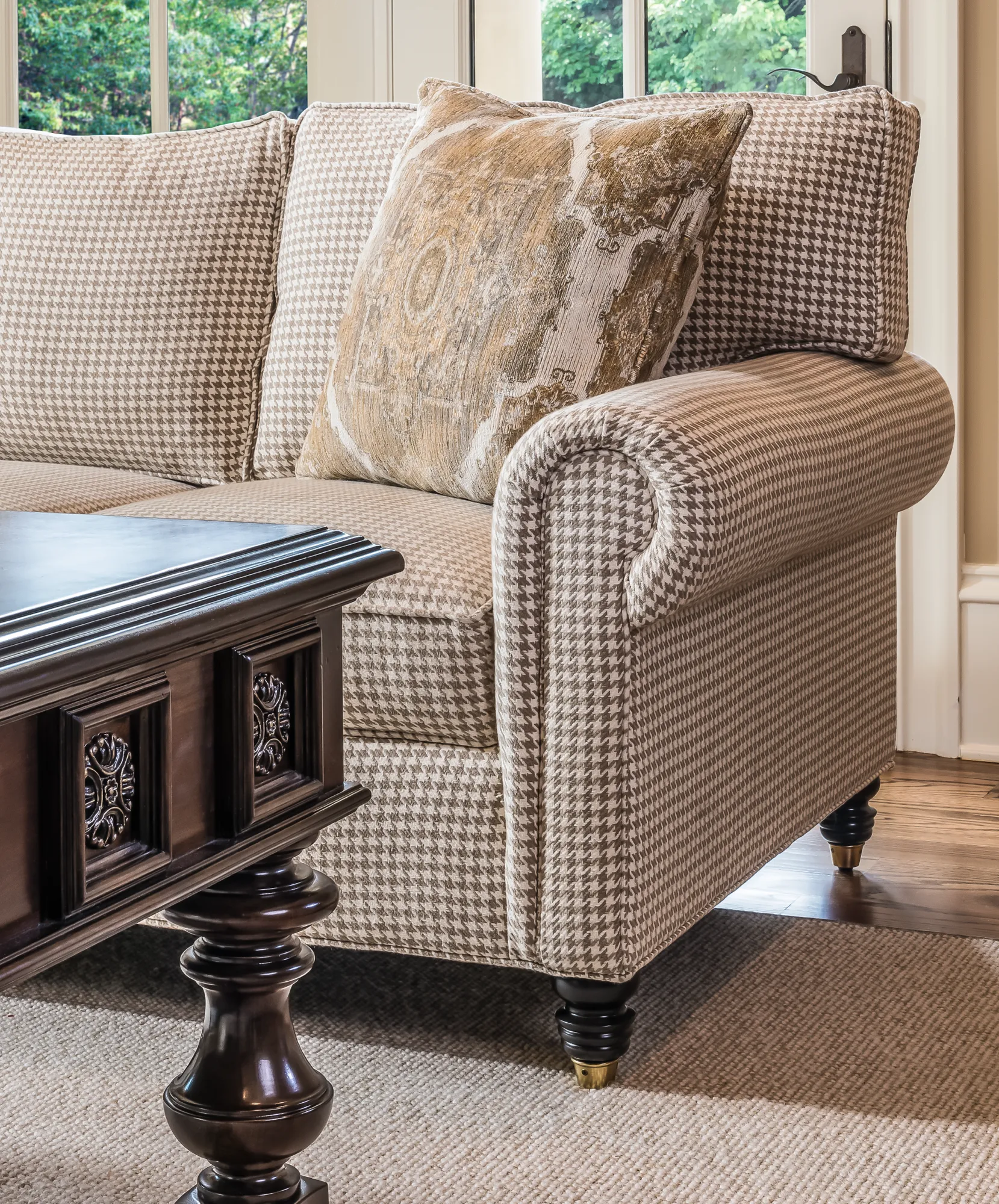 A traditional sofa upholstered in tan houndstooth fabric with a gold accent pillow. In the foreground is a dark, carved wood table, with green trees visible through the window.
