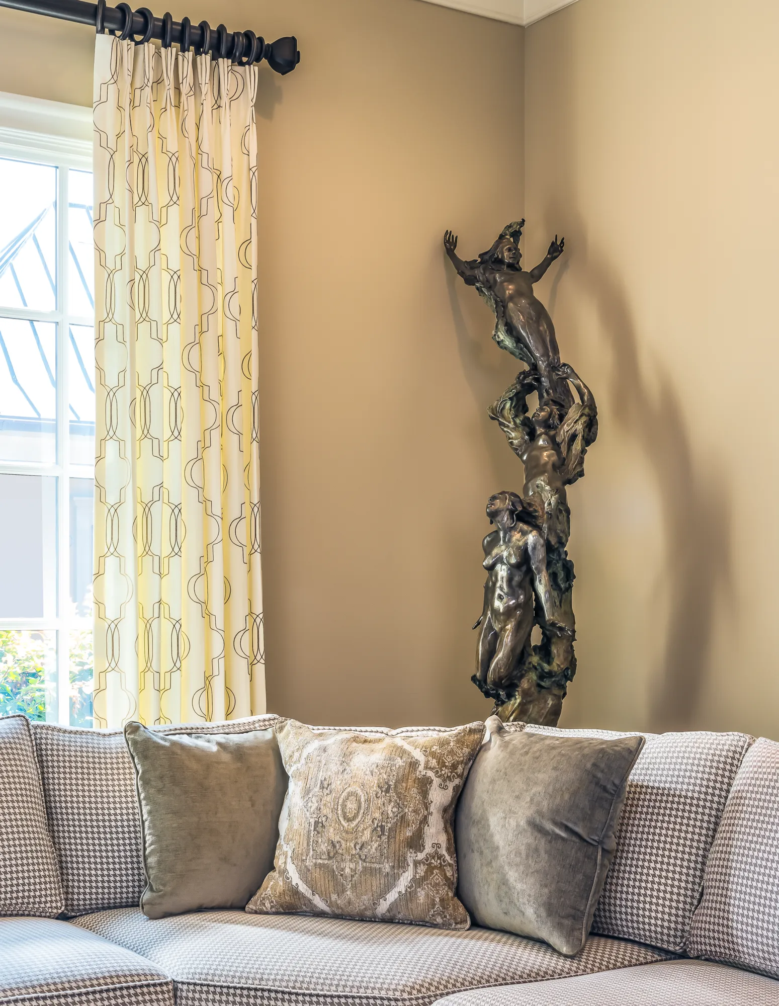 A gray patterned sofa with plush pillows is shown in the foreground. A tall, bronze sculpture of intertwined figures accents the corner next to a geometric-patterned curtain.