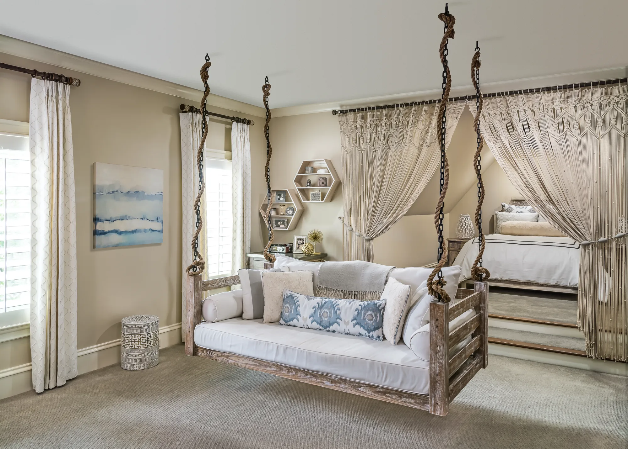 A weathered wood hanging daybed with white cushions and blue pillows suspended by thick ropes in a neutral bedroom with a raised sleeping nook covered by macrame curtains.
