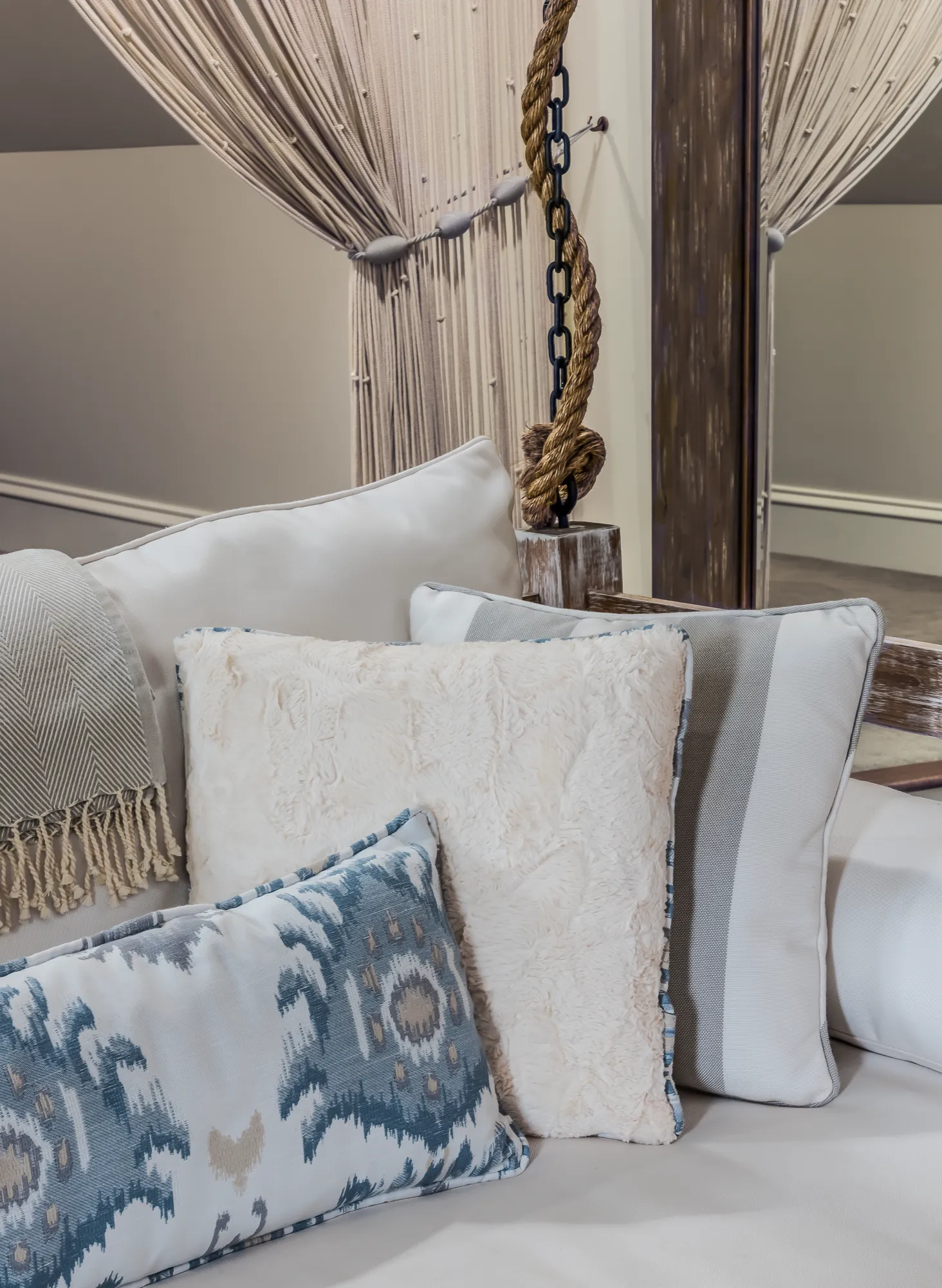 A grouping of textured cream, gray, and blue decorative pillows and a throw blanket arranged on a sofa. A thick rope, chain, and macrame string curtain are visible in the background.