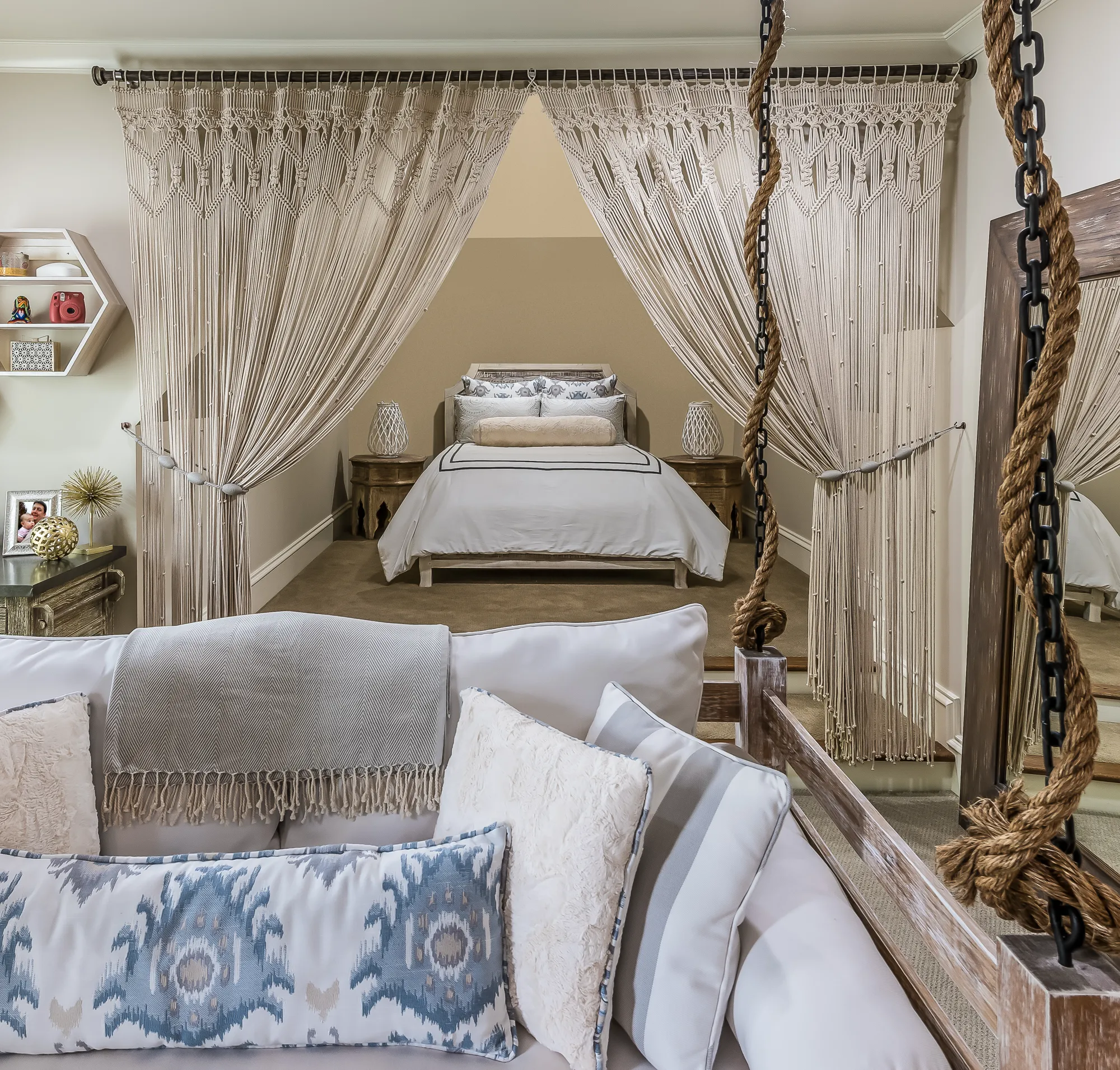 A plush swing seat with pillows sits before a bedroom area framed by dramatic, tied-back macrame string curtains, revealing a bed with white linens.