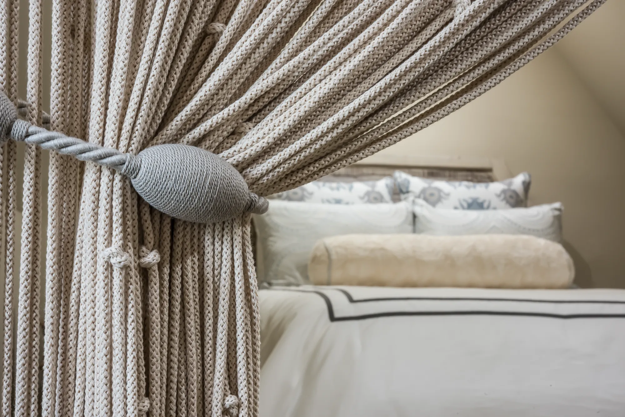 A beige, woven rope curtain is pulled aside by a decorative gray tie-back, framing a view of a neatly made bed with neutral white and patterned pillows.