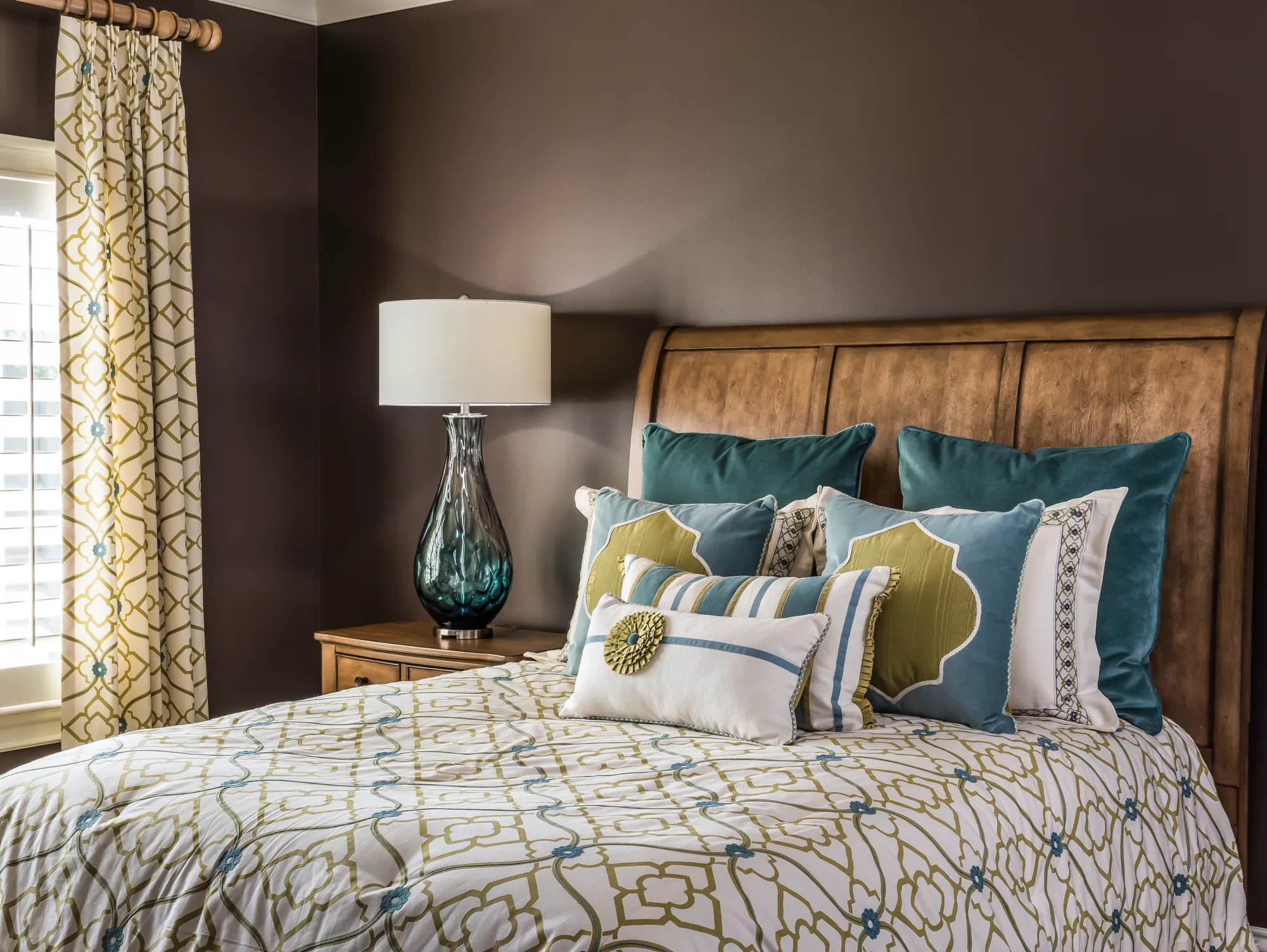 A richly decorated bedroom showcases a large wooden headboard, dark brown walls, and a patterned duvet. Numerous decorative pillows in teal and green complete the look.