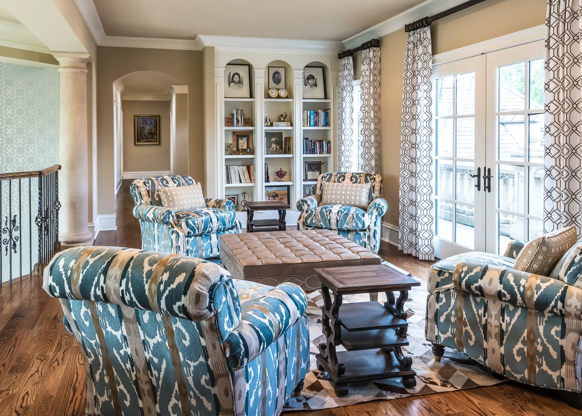 A comfortable sitting room featuring four patterned blue chairs, a leather ottoman, built-in bookcases, and French doors leading to a bright exterior.