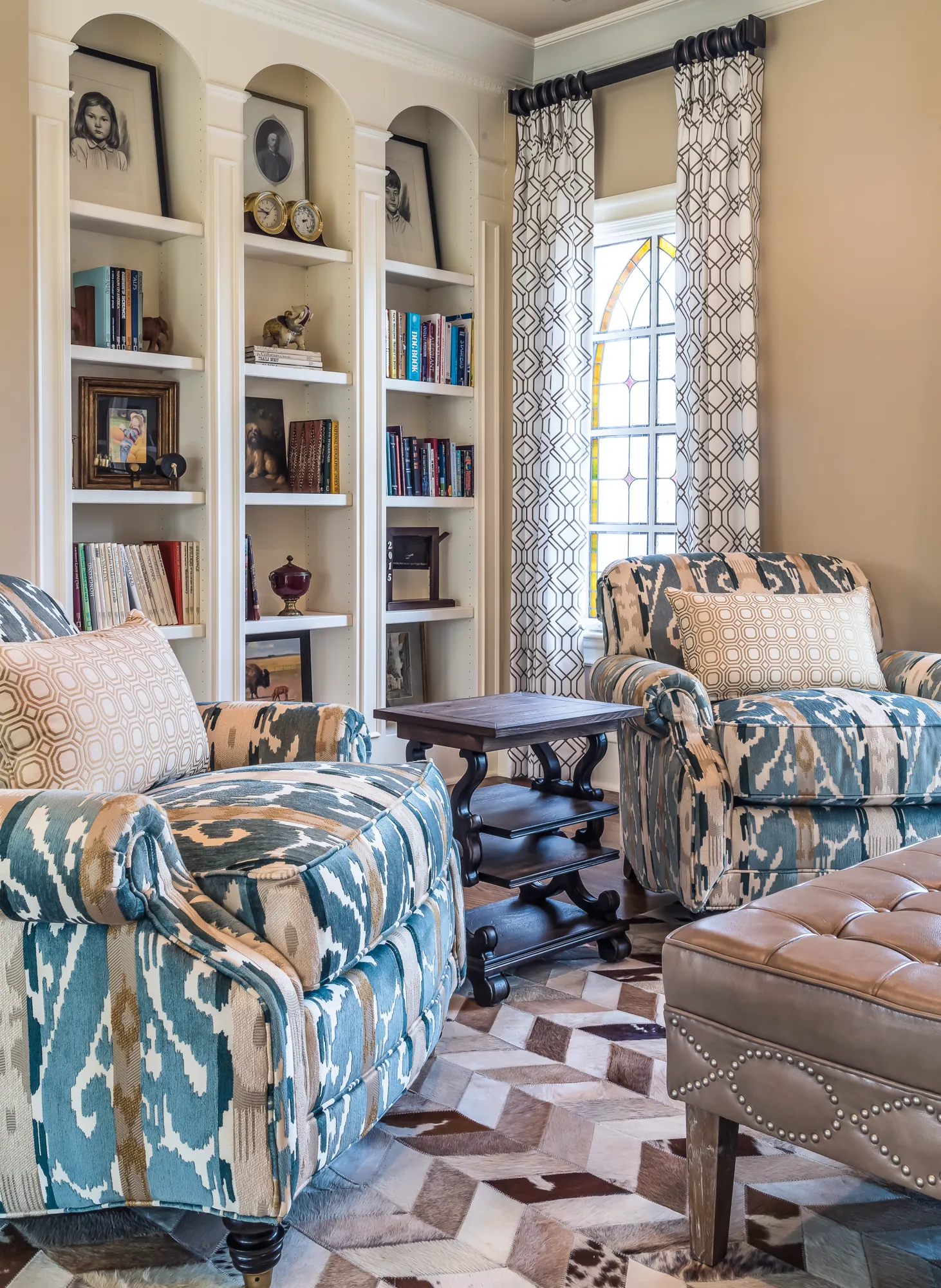 A den with custom white built-in bookshelves displaying books and framed photos. Two blue patterned armchairs and a leather ottoman rest on a geometric rug.