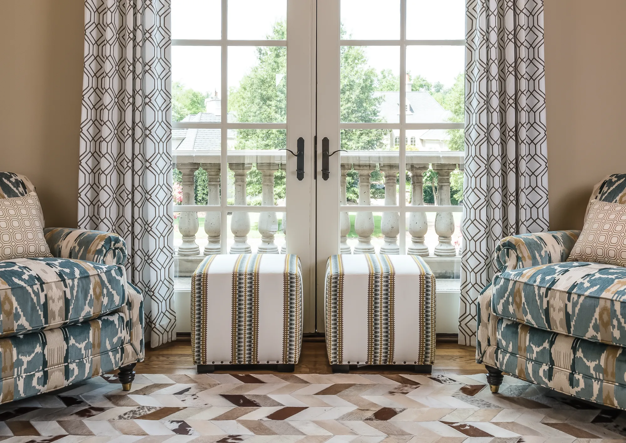 A luxurious seating area featuring two patterned armchairs, two striped ottomans, and a chevron hide rug framing white French doors that open onto a stone balustrade balcony.