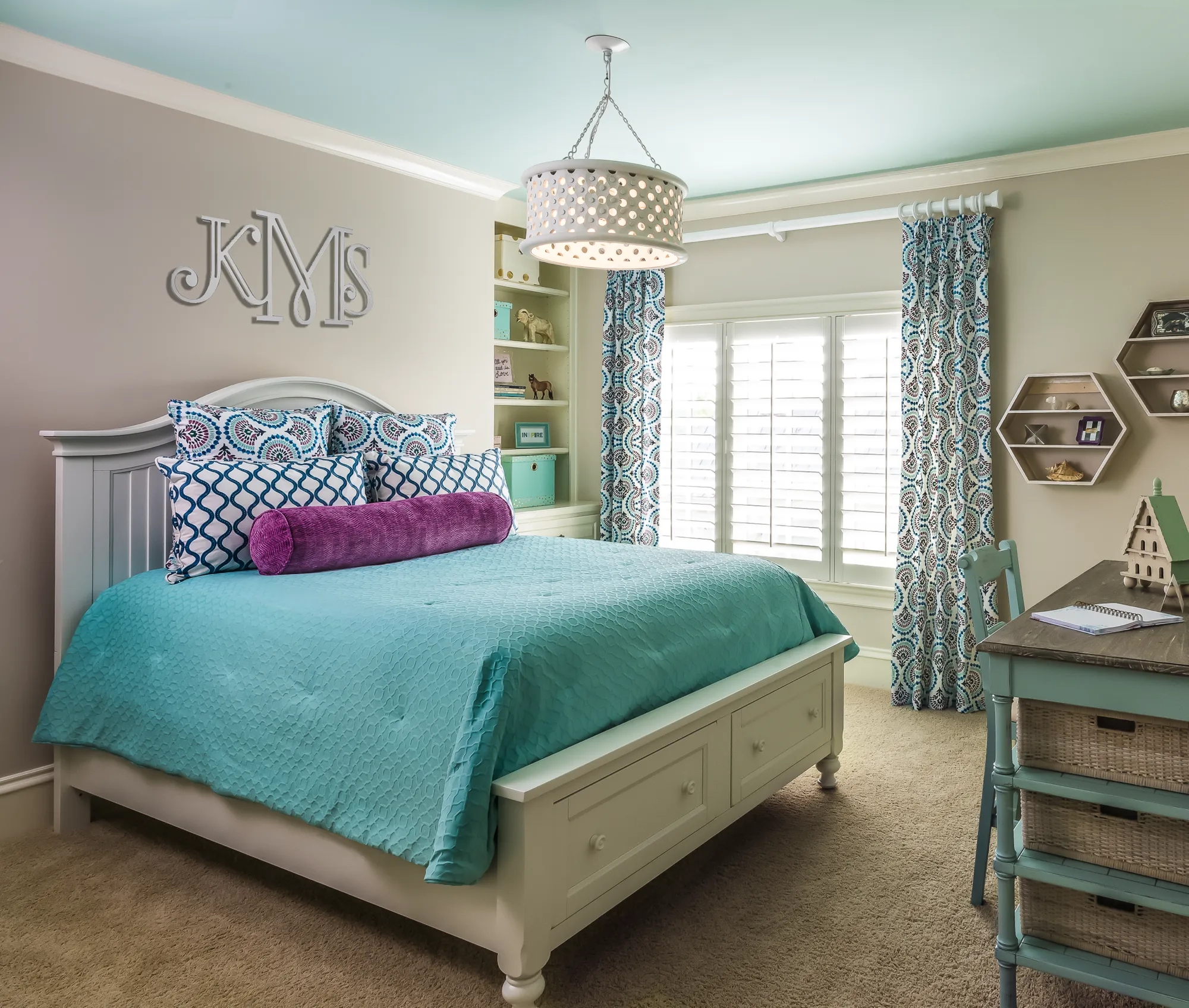 A vibrant, custom-designed bedroom with a white bed frame, turquoise bedding, and a purple bolster pillow. The pale blue ceiling complements the patterned curtains and wall monogram.