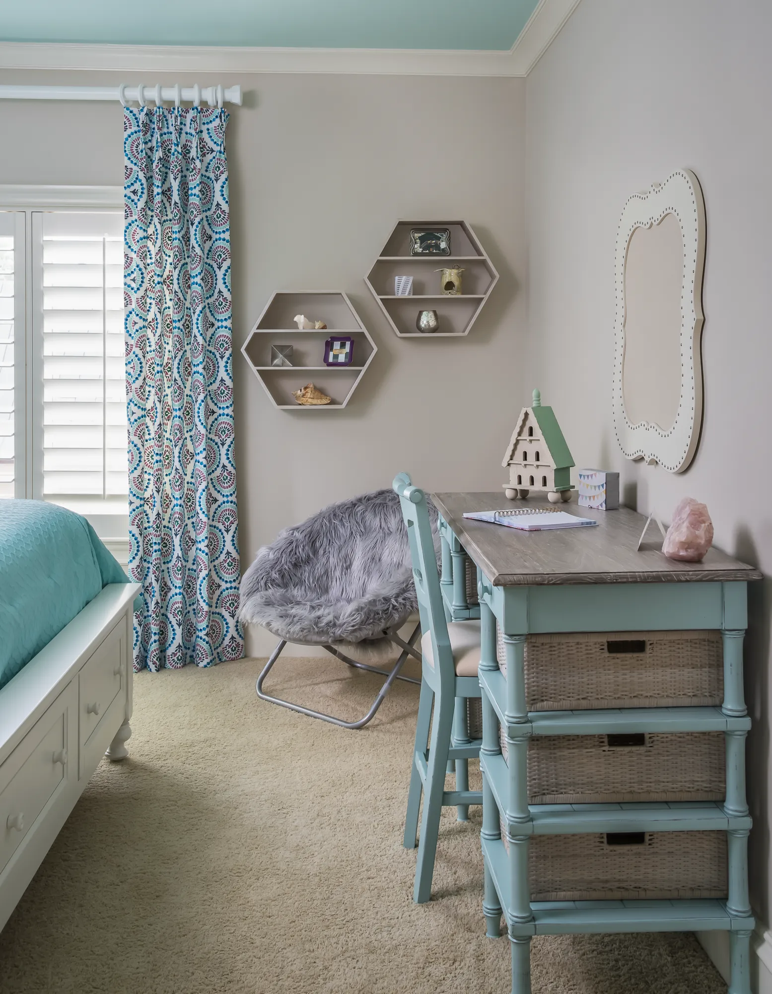 A bright bedroom corner featuring a teal-painted desk with wicker drawers, a matching chair, and a white bed with a turquoise spread. Hexagonal wall shelves hold trinkets.