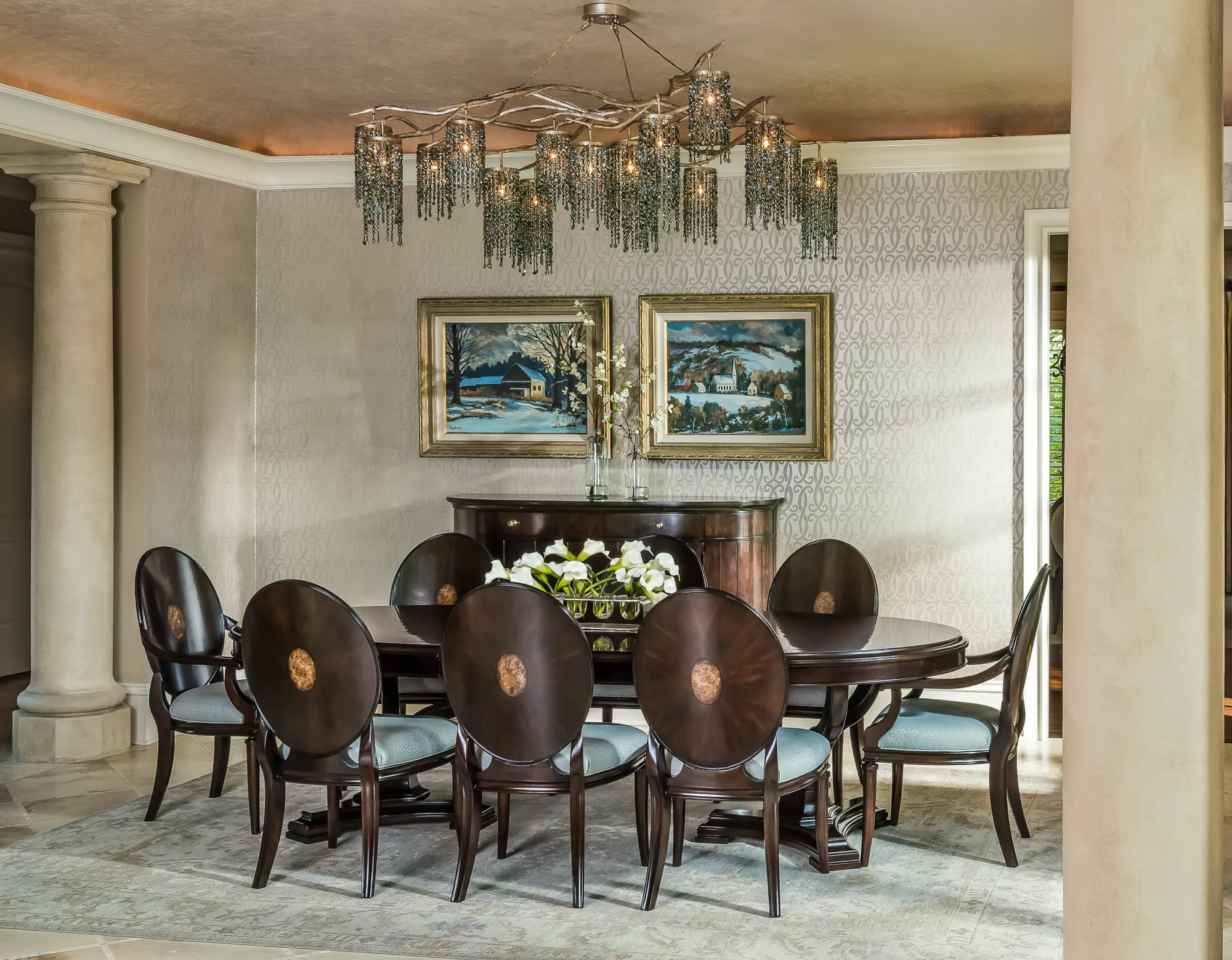 An elegant dining room features a dark oval table and eight high-backed chairs with unique wood inlays. A crystal branch chandelier hangs over a centerpiece of white calla lilies.