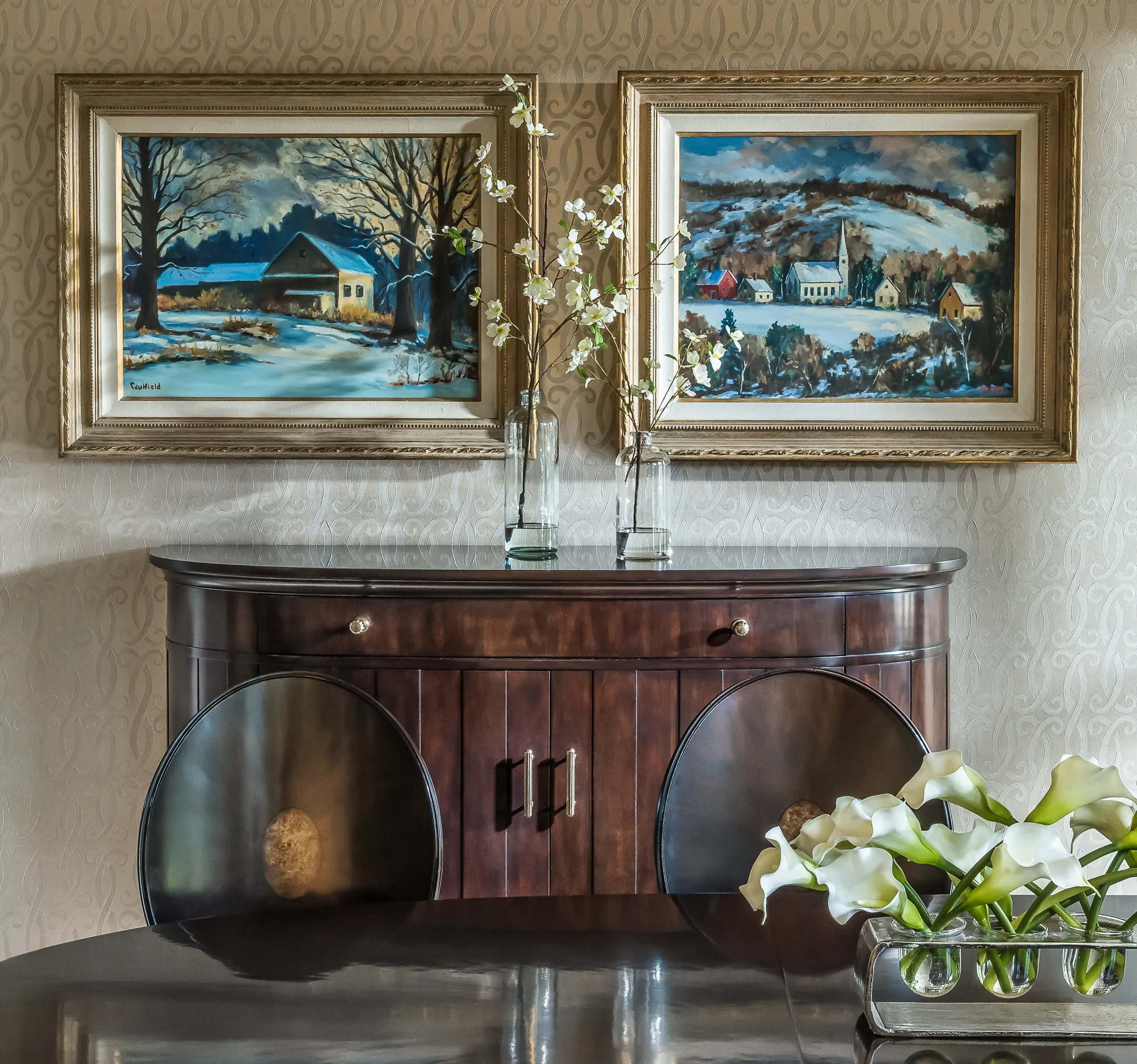Two framed winter landscape paintings are centered above a dark, curved credenza. White flowers in glass vases sit on the credenza and the polished dark dining table in the foreground.