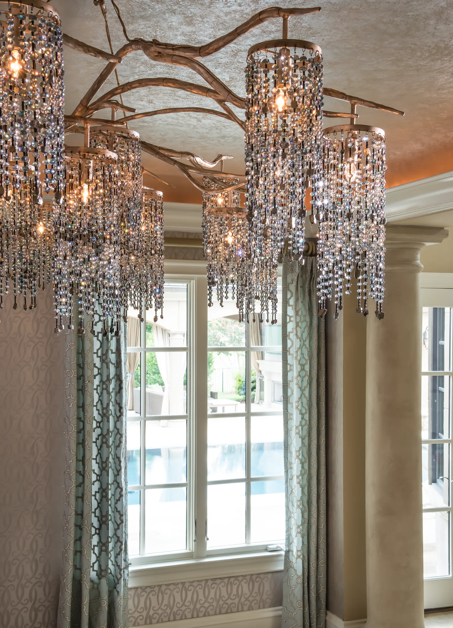 A unique, multi-cylinder crystal chandelier, suspended by metallic branches, illuminates a room with patterned wallpaper and drapes framing a large window.