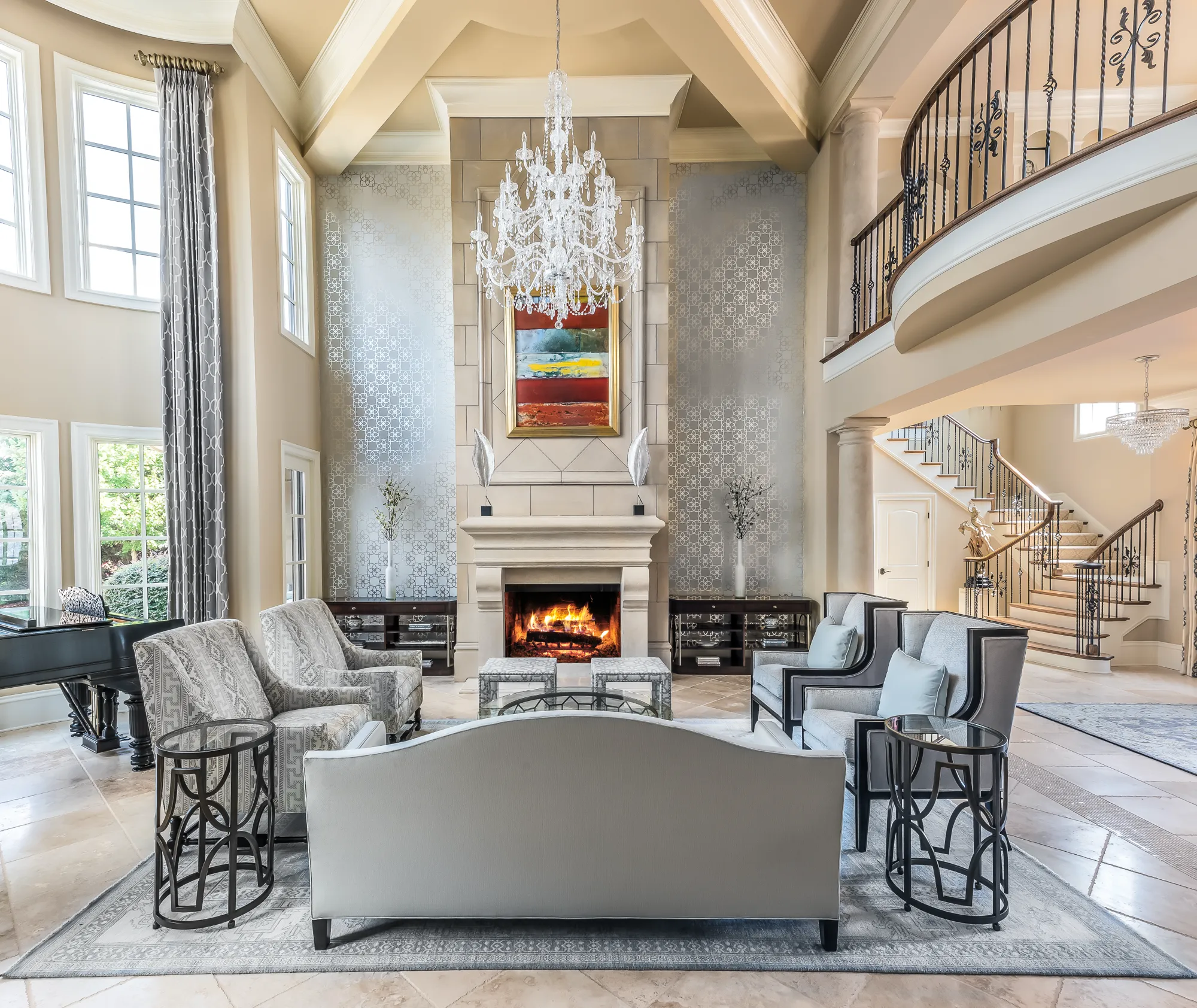 A luxurious two-story living room is centered on a stone fireplace below a chandelier and abstract art. The space features formal seating, a grand piano, and a curved staircase leading to the second-floor balcony.