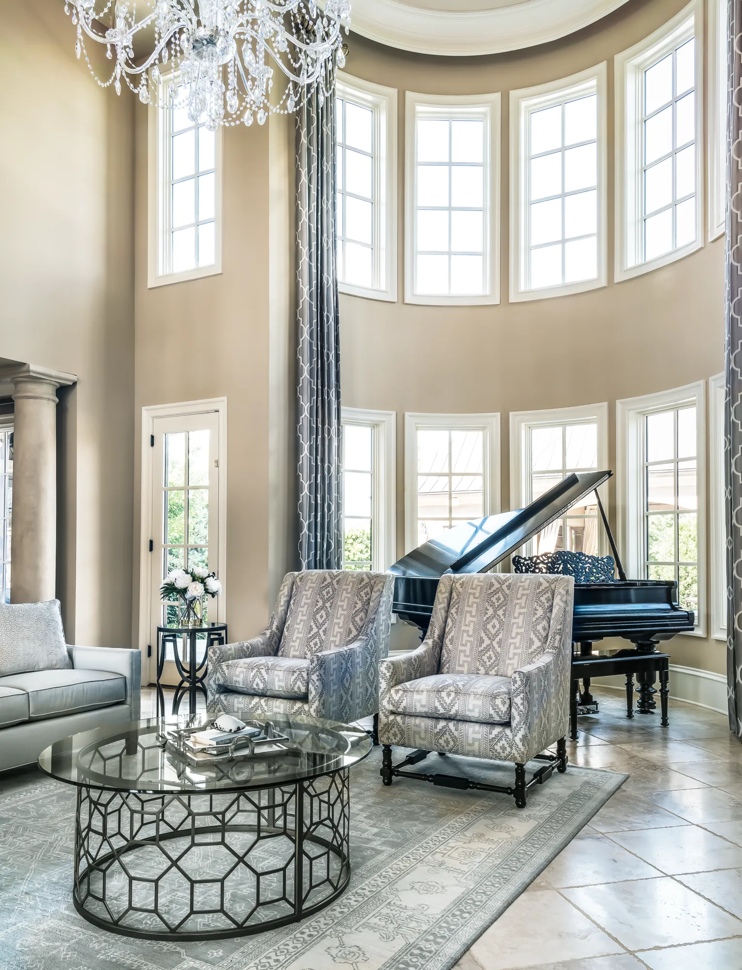 A grand living room featuring a tall, curved wall of windows, a crystal chandelier, and a shiny black grand piano. Two patterned armchairs frame a glass coffee table.