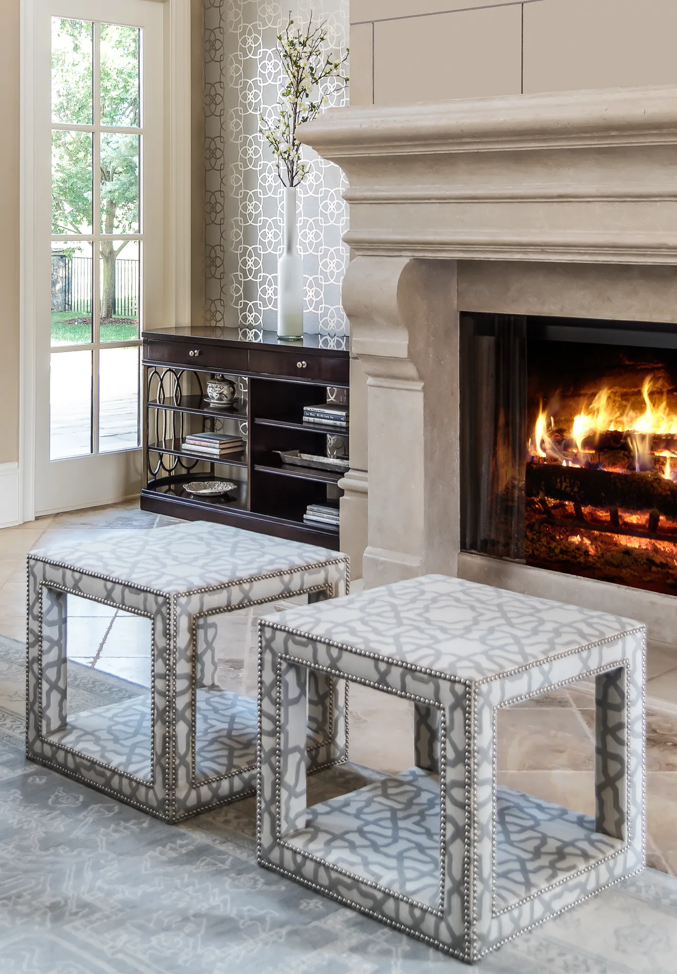 A sophisticated living space featuring a lit stone fireplace and two grey patterned upholstered ottomans set on a light rug, next to a dark console table and glass French doors.