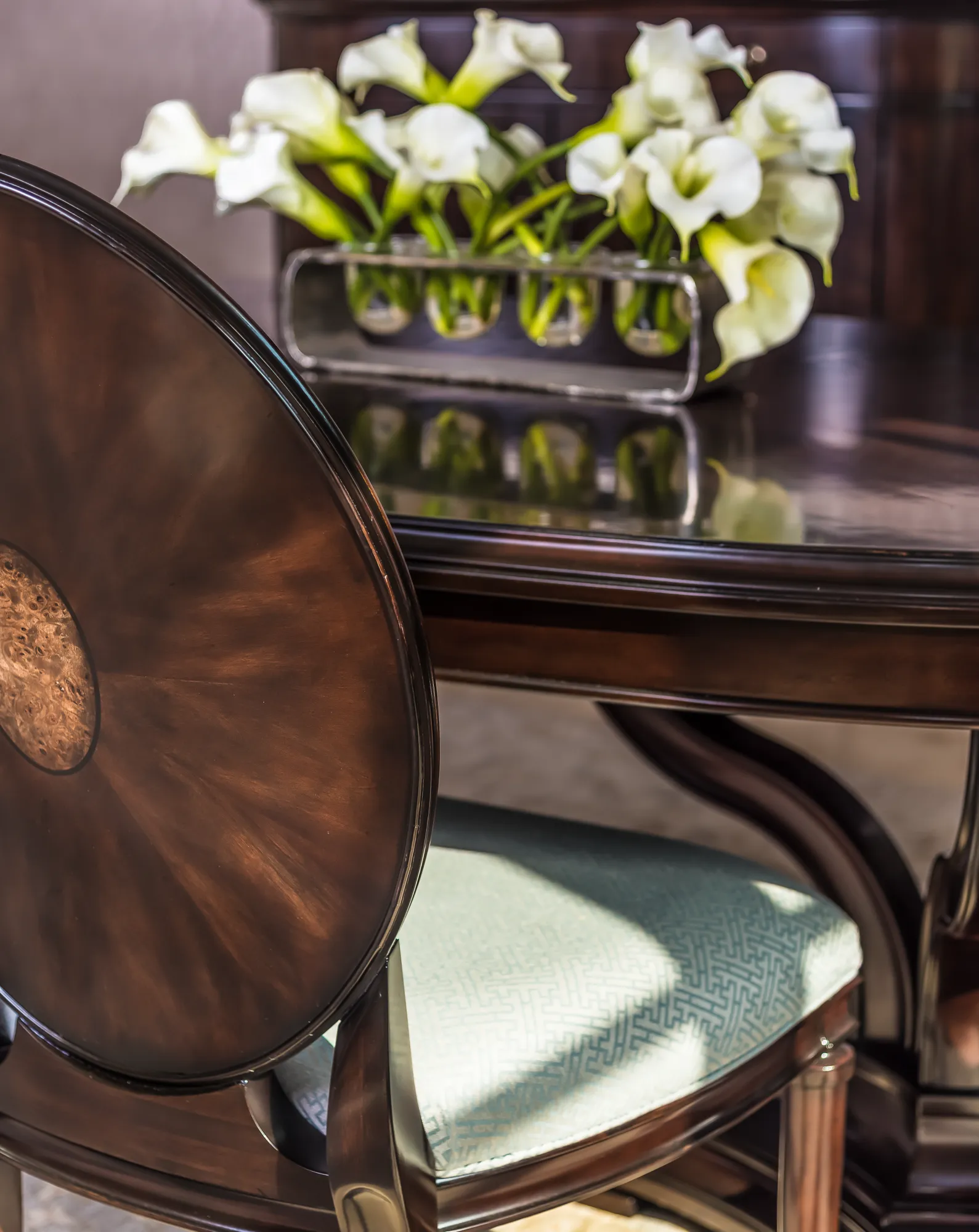 Dark, polished wood furniture, featuring a chair and table topped with white calla lilies in a sleek, silver vase.