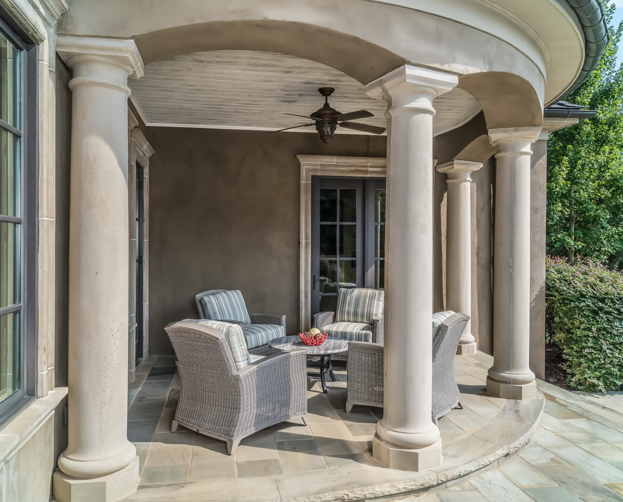 A curved, covered outdoor patio features classic columns, dark stucco walls, and a ceiling fan. Four comfortable wicker chairs with striped cushions sit around a small round table.