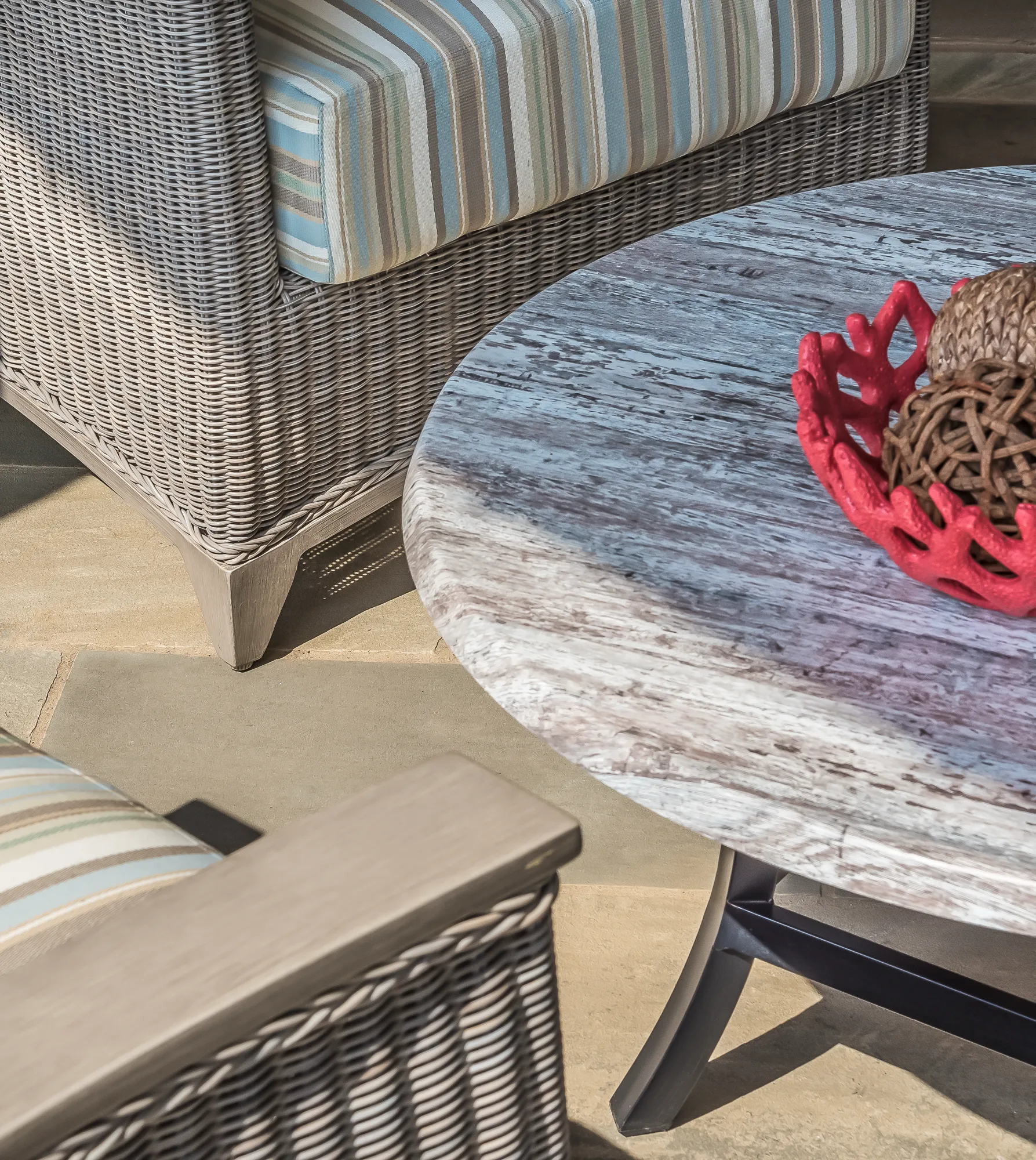 Close-up of outdoor furniture: light gray wicker chairs with striped cushions flank a round, weathered wood table decorated with a red coral bowl.