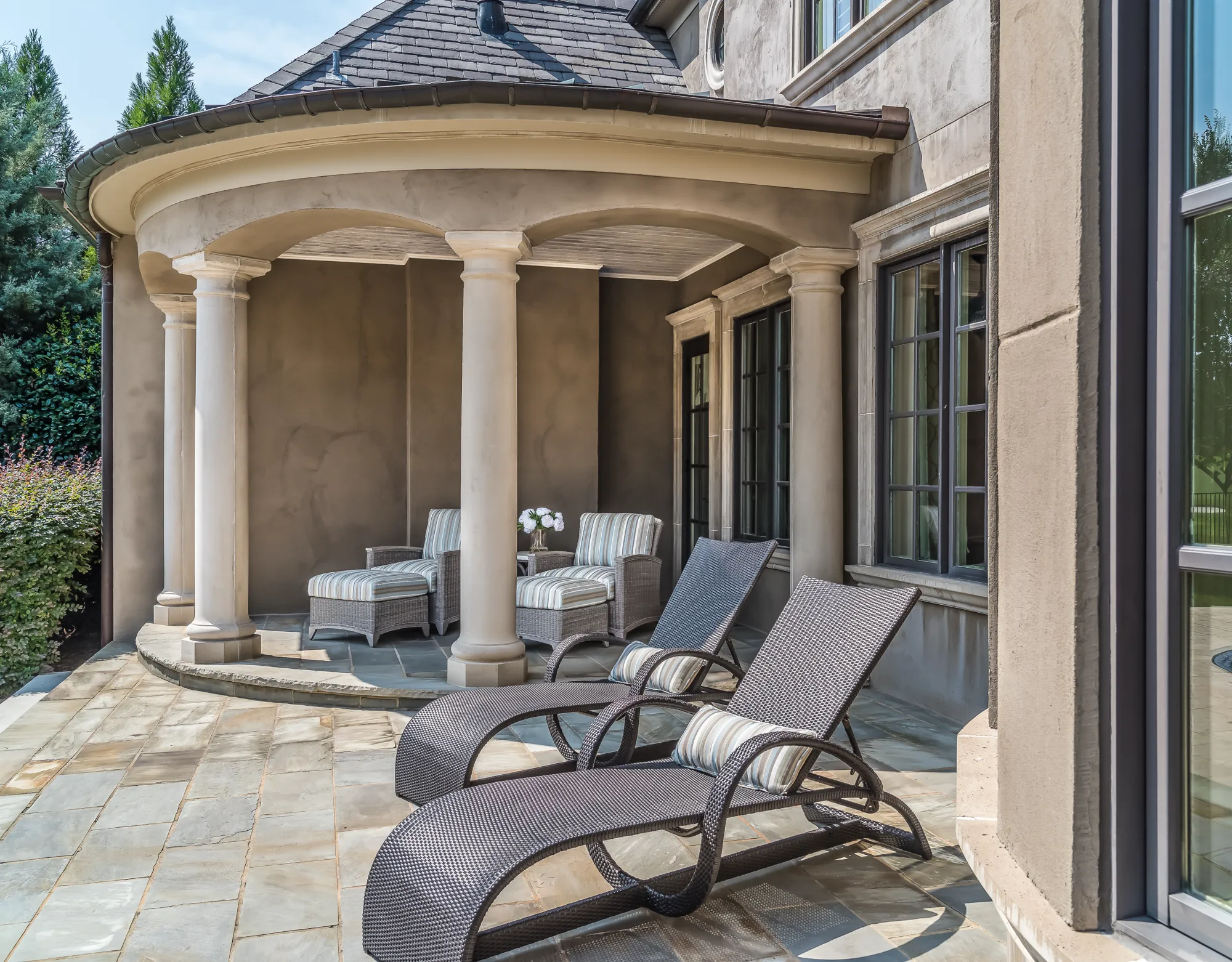 A luxurious outdoor terrace with a stone floor and covered portico features two wicker lounge chairs and two cushioned armchairs.