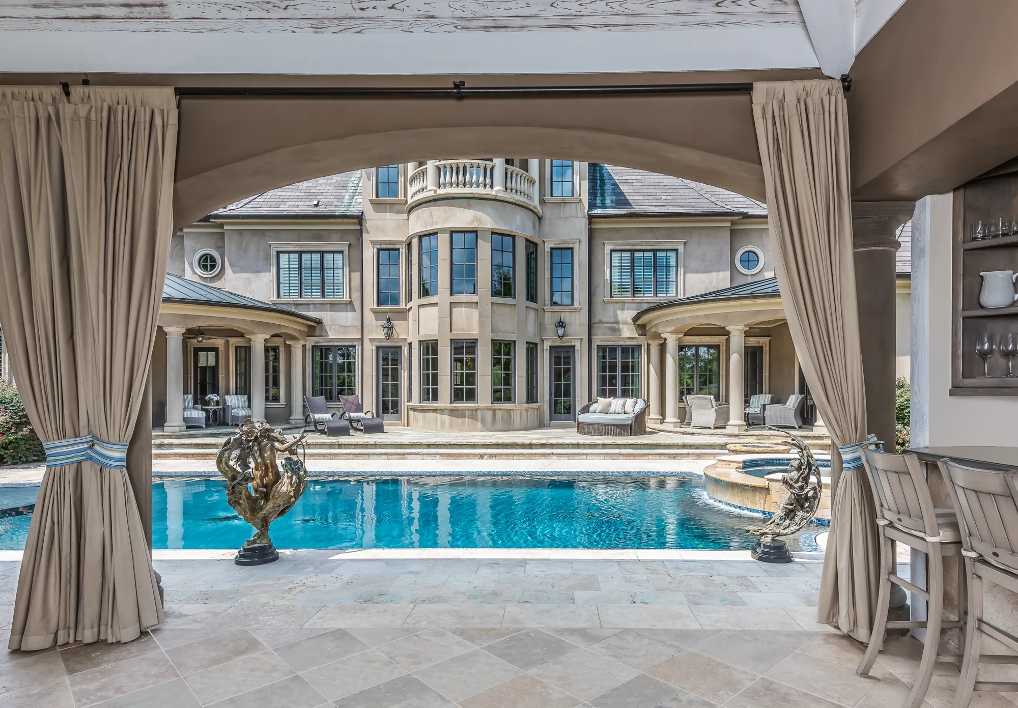The view from an outdoor cabana, framed by beige curtains, features a bright blue pool and a stone patio leading to the classic, multi-story exterior of a luxury home.
