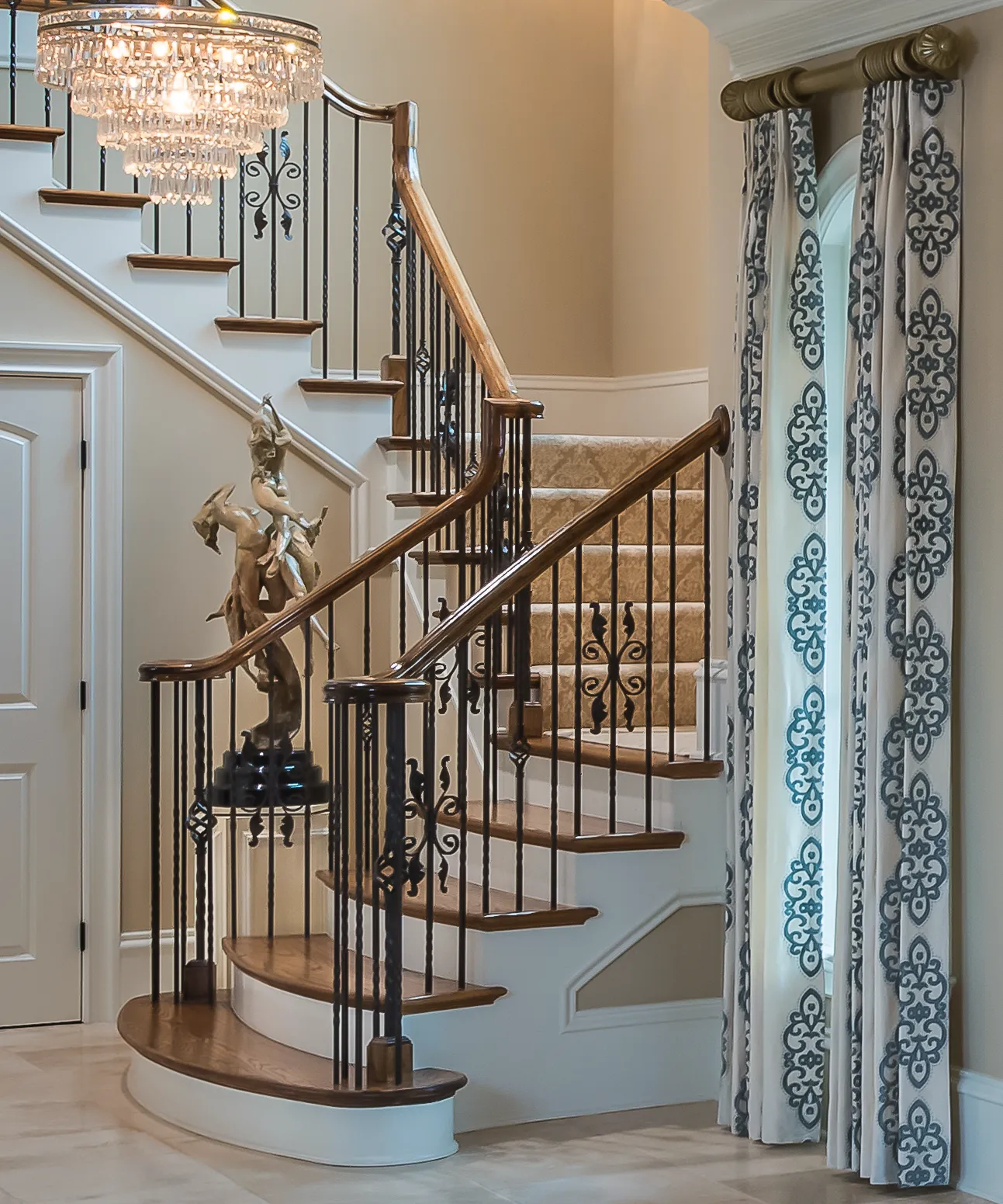 An elegant, curved staircase with a wood and iron railing, carpet runner, and a classical sculpture on the newel post. A crystal chandelier illuminates the foyer.