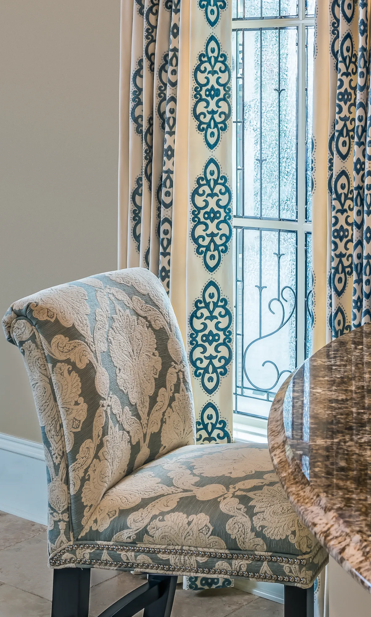 A luxury interior detail showing an upholstered chair with blue damask next to a polished granite counter. The window features textured glass and custom drapes with a matching blue pattern.