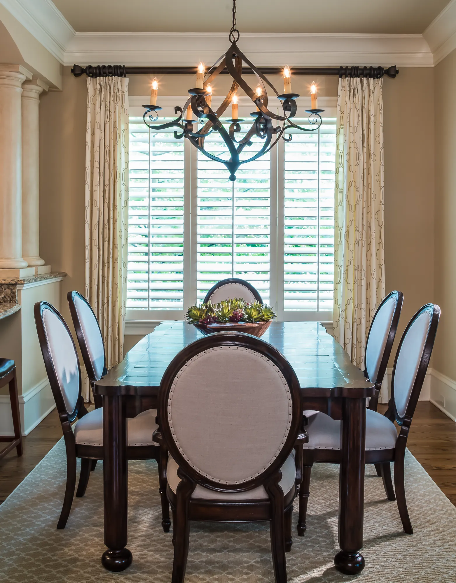 A traditional dining room featuring a dark wood table and six cream upholstered chairs on a patterned rug. An ornate iron chandelier is centered above the table, set against a window with white shutters.