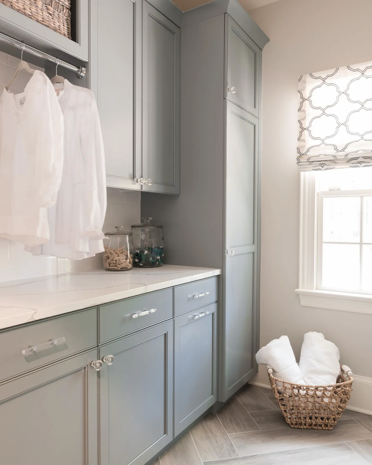 A bright, modern laundry room featuring soft gray cabinets, white quartz countertops, and decorative glass knobs. White shirts hang above the counter, and fresh white towels are in a woven basket next to a window.