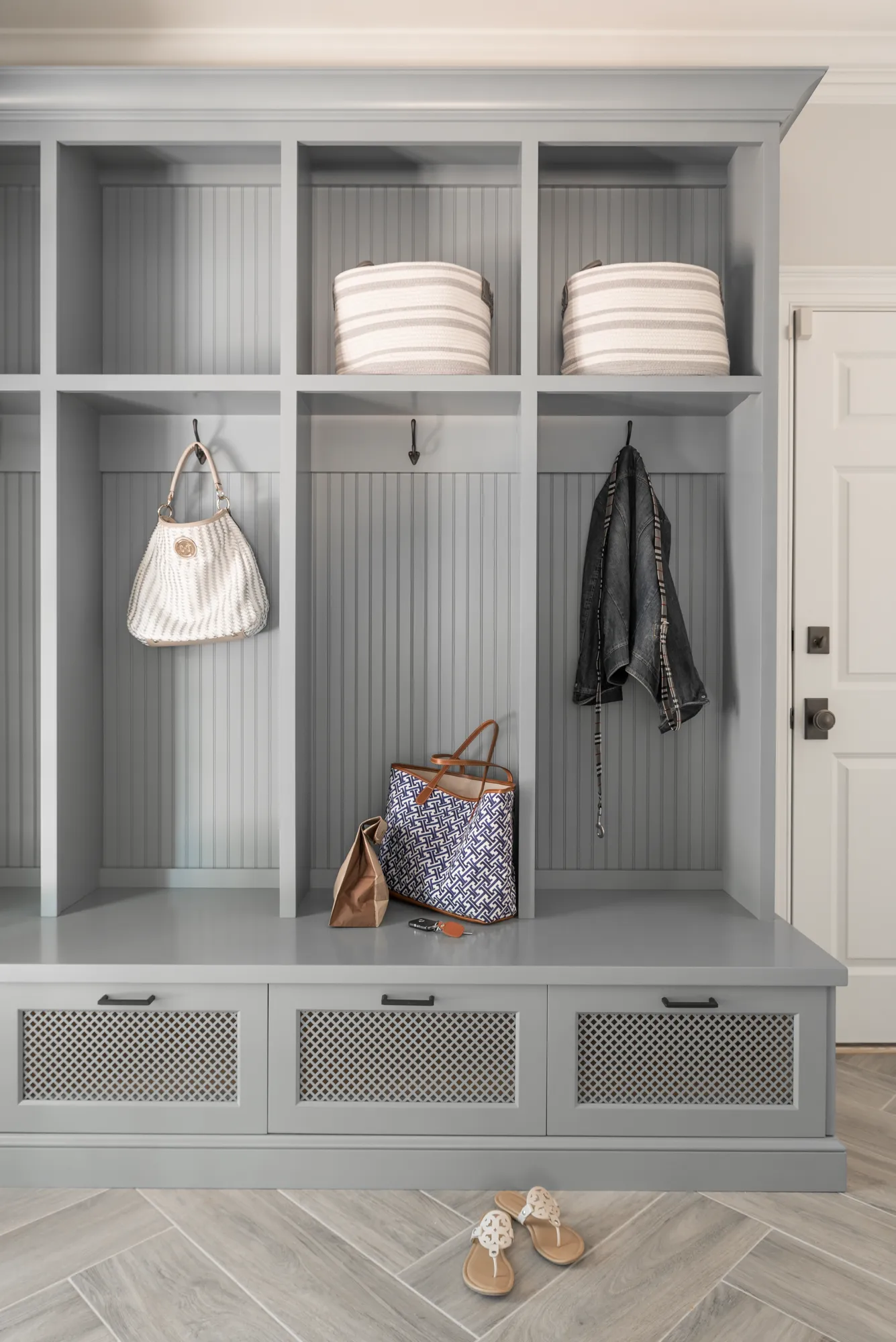 A custom light gray mudroom unit with beadboard backing and lattice-front drawers. The unit holds woven baskets, a jacket, keys, and various bags above a herringbone tile floor.
