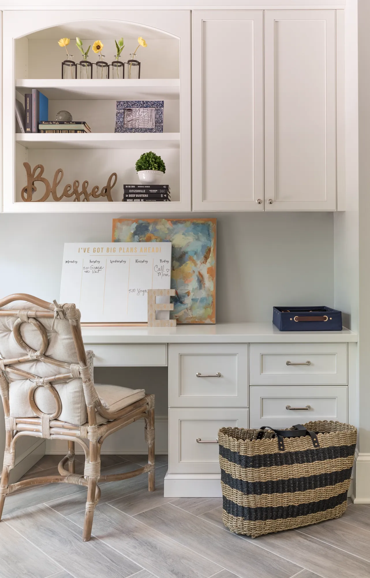 A clean, bright home office nook with white built-in cabinetry and drawers. A rattan chair sits before the desk, which is decorated with a planner and abstract art.