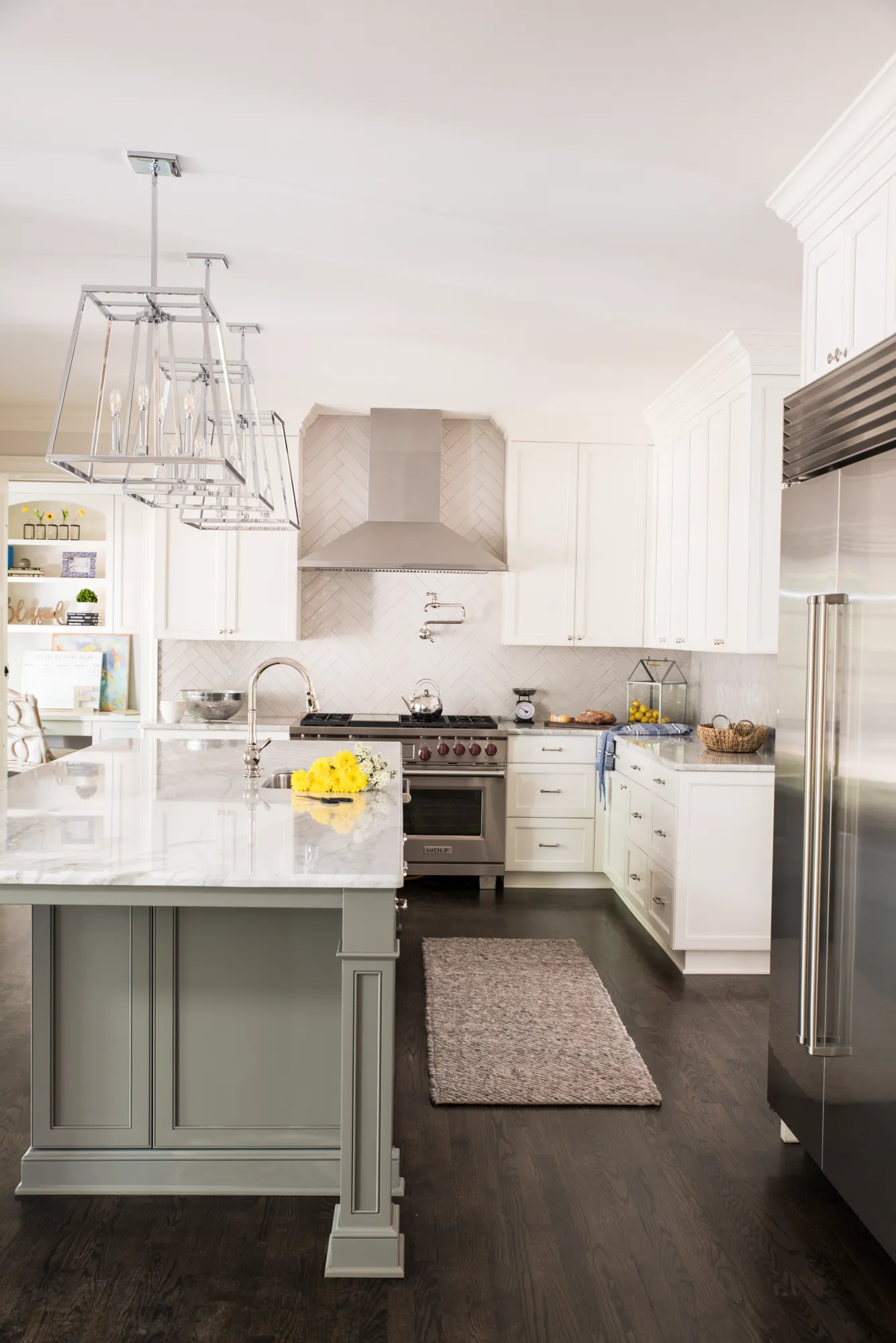A bright, modern kitchen featuring white shaker cabinets, dark floors, and stainless steel appliances. A large island with a sage base and marble counter sits below chrome light fixtures.