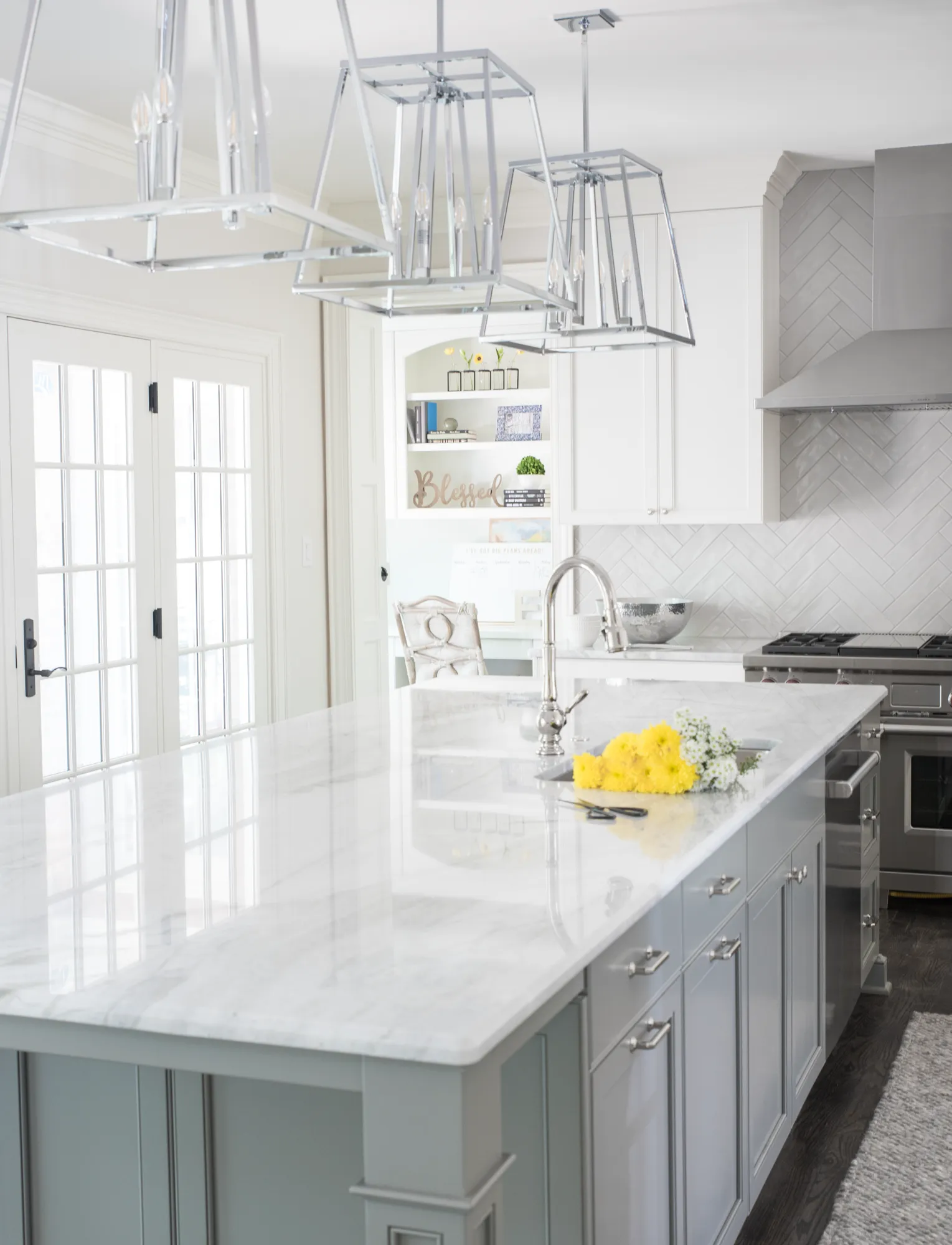 A bright kitchen showcasing a large island topped with white marble, illuminated by chrome pendant lights. The background features white cabinets and a light gray herringbone tile backsplash.