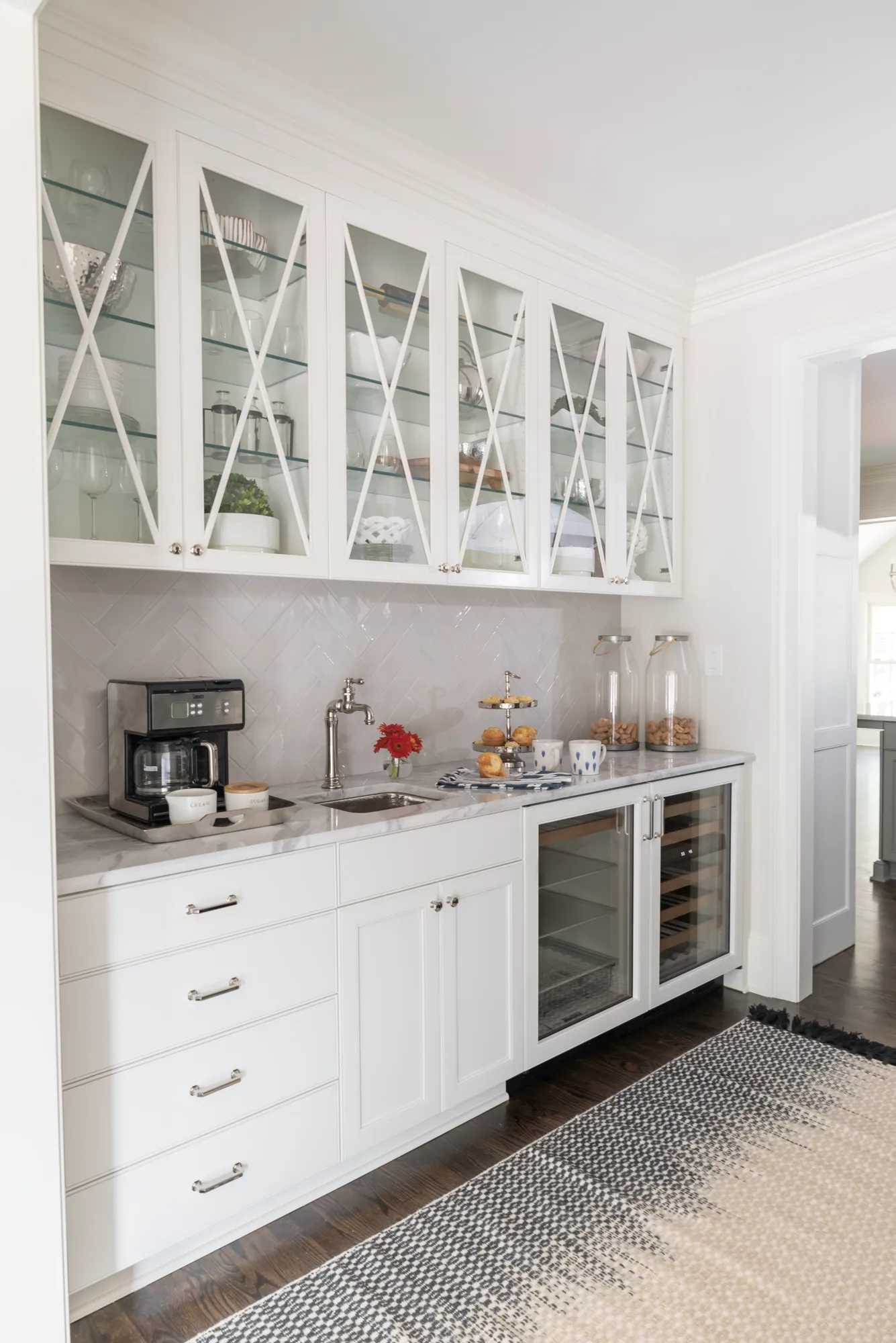 A custom white wet bar with marble counters, a herringbone backsplash, and crisscrossed glass upper cabinets. The bar features a sink, coffee maker, small pastries, and an undercounter beverage fridge.