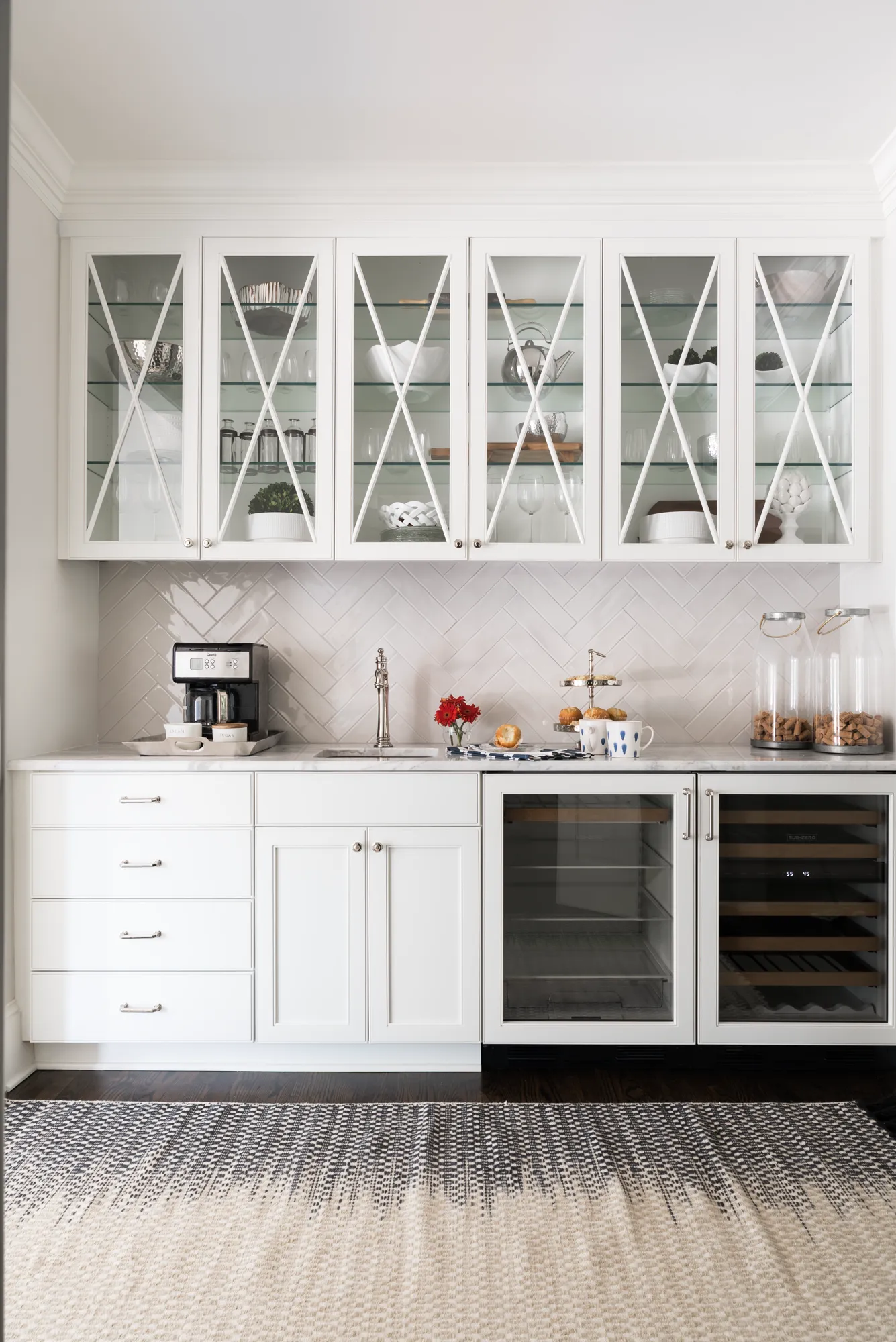 A luxurious custom white wet bar featuring glass X-mullion upper cabinets, white herringbone tile, and a marble counter. It includes a coffee maker, sink, and dual under-counter refrigeration units.