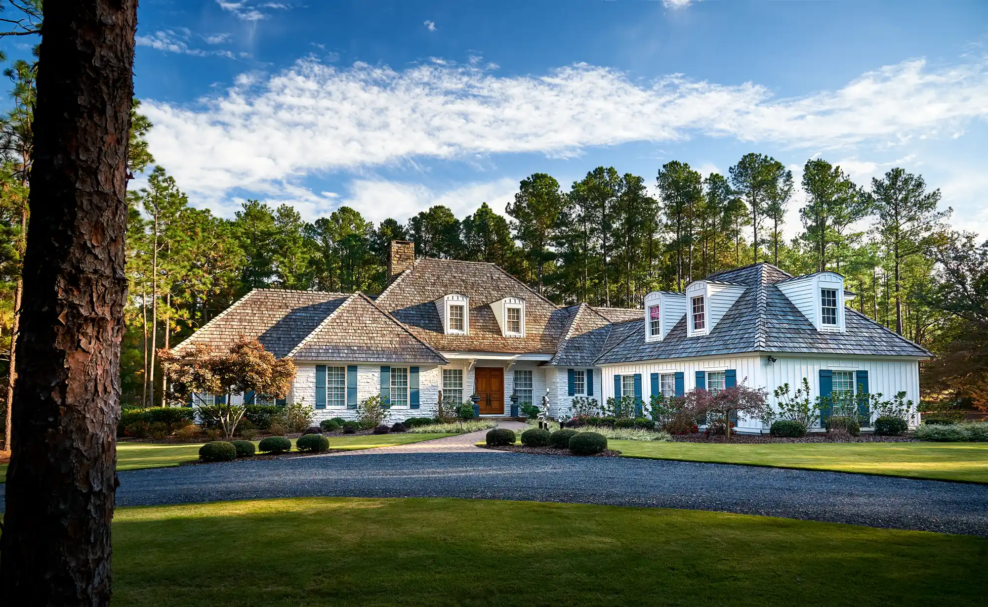 A beautiful white stone house with dark wooden shingles and teal shutters stands on a green lawn. A gravel driveway leads to the entrance, framed by a thick forest of tall pine trees.