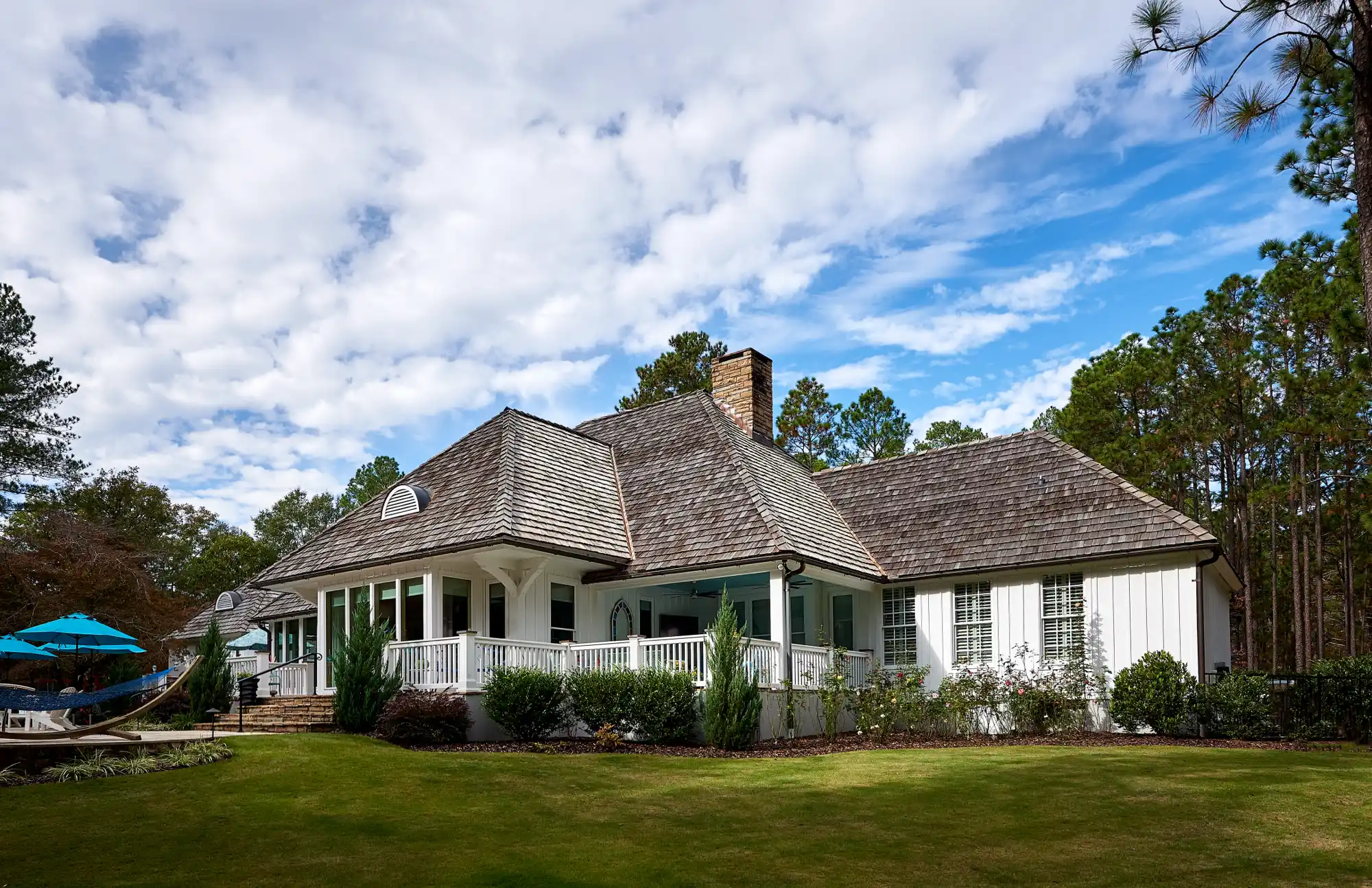 A luxury white ranch home with a shingle roof and a stone chimney overlooks a groomed green lawn, surrounded by a dense forest of tall pine trees.