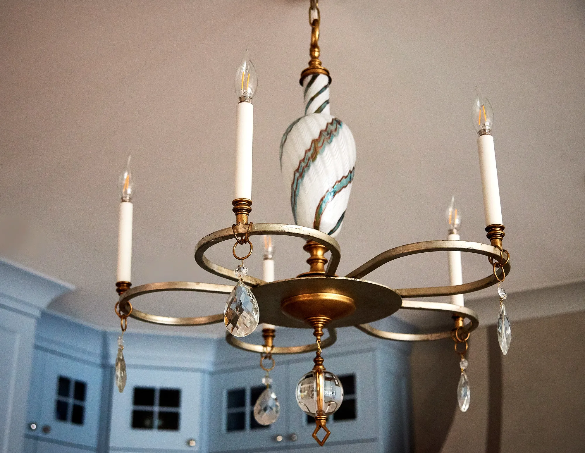 A brushed gold candelabra chandelier hangs from the ceiling, featuring crystal teardrops and a central white glass piece with teal swirls. Blue cabinetry is visible below.