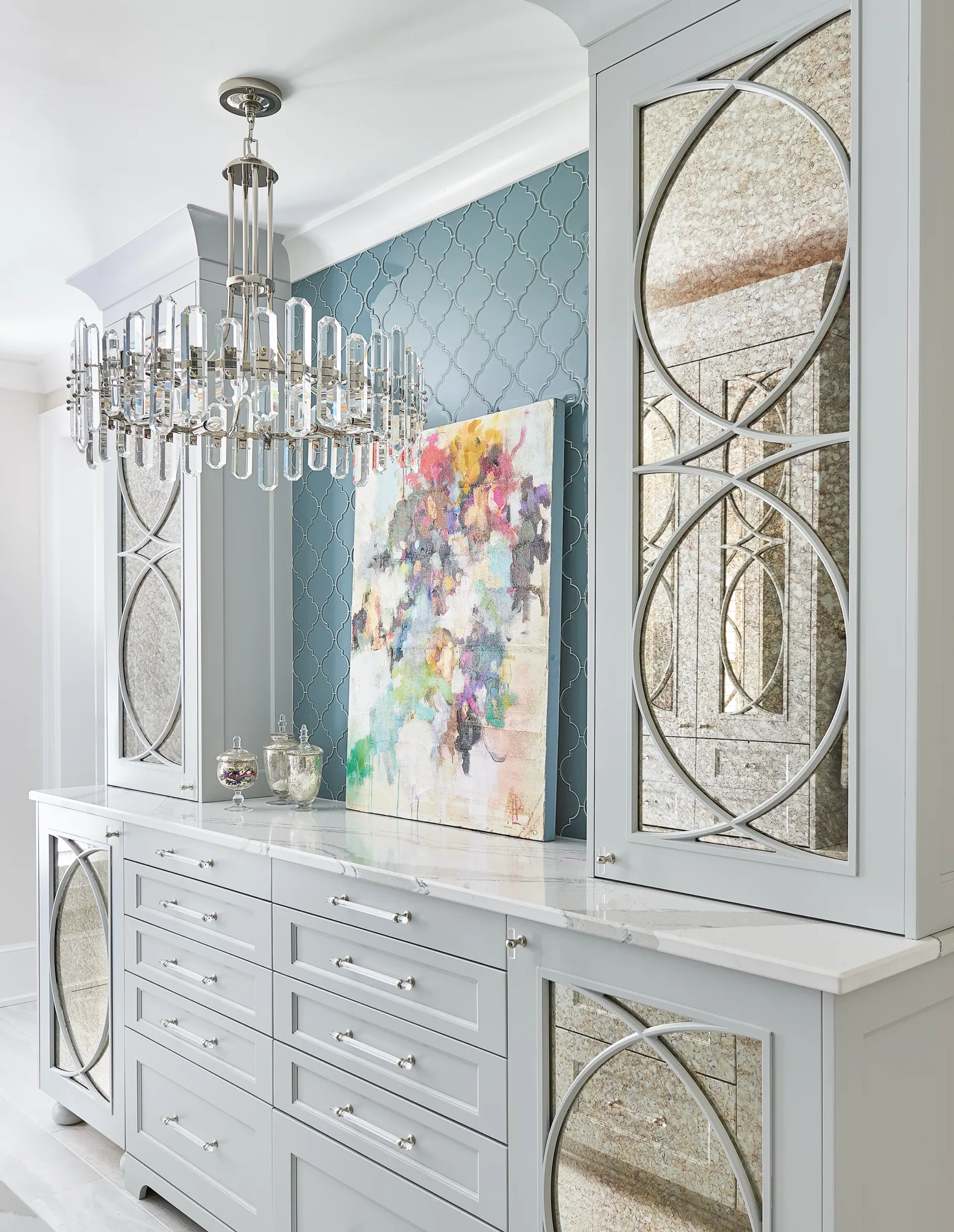 A modern crystal chandelier illuminates a gray custom vanity with a marble top and decorative mirrored doors. A colorful abstract painting rests against blue arabesque tile.