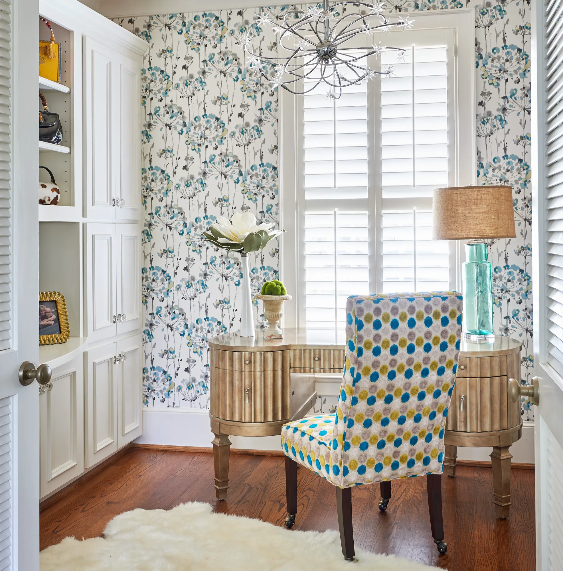 A stylish home office with floral wallpaper, white built-in storage, and a gold desk facing a shuttered window. A colorful polka-dot chair is centered on a white rug.