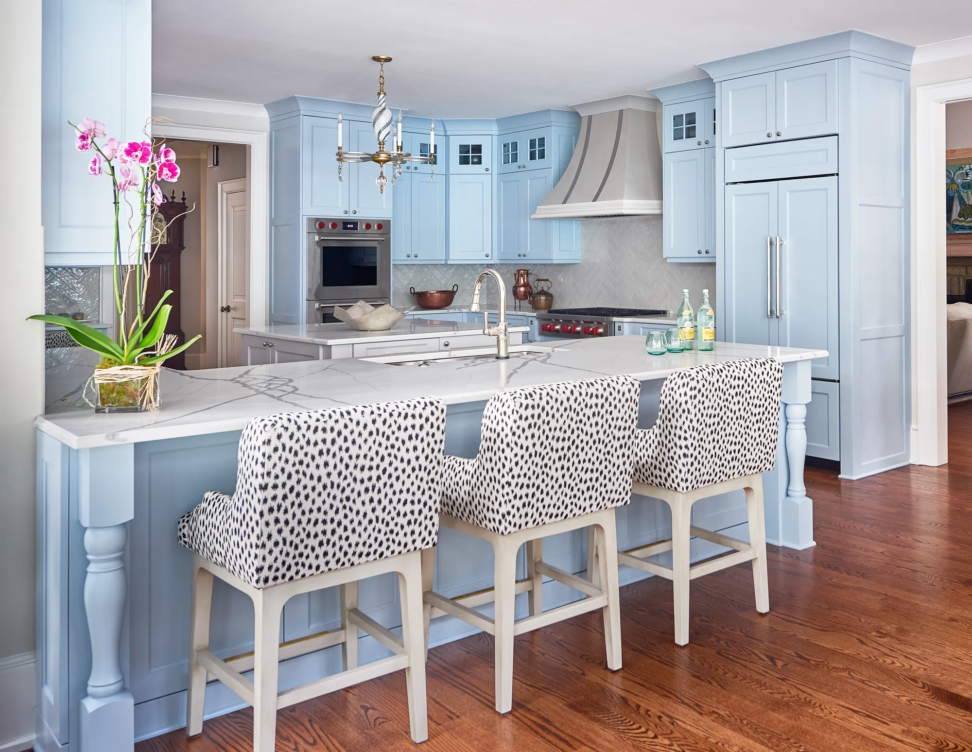 A bright kitchen with pale blue cabinets, a marble island, and hardwood floors. Three barstools with black and white spotted upholstery are pulled up to the island.