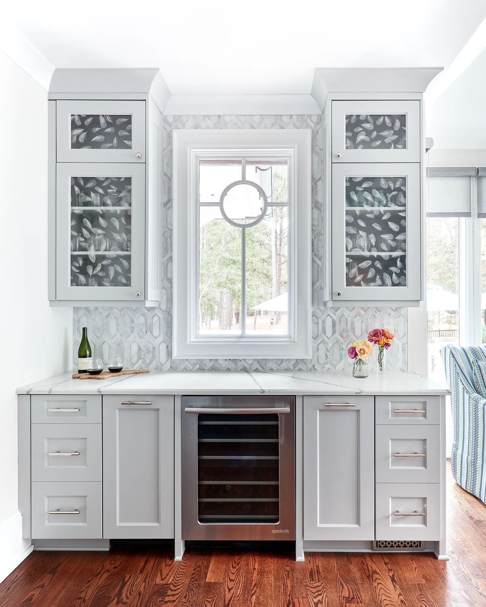 A custom home beverage bar features light gray shaker cabinets, a marble countertop, and a built-in wine refrigerator below a window. The marble backsplash has an intricate patterned tile design.