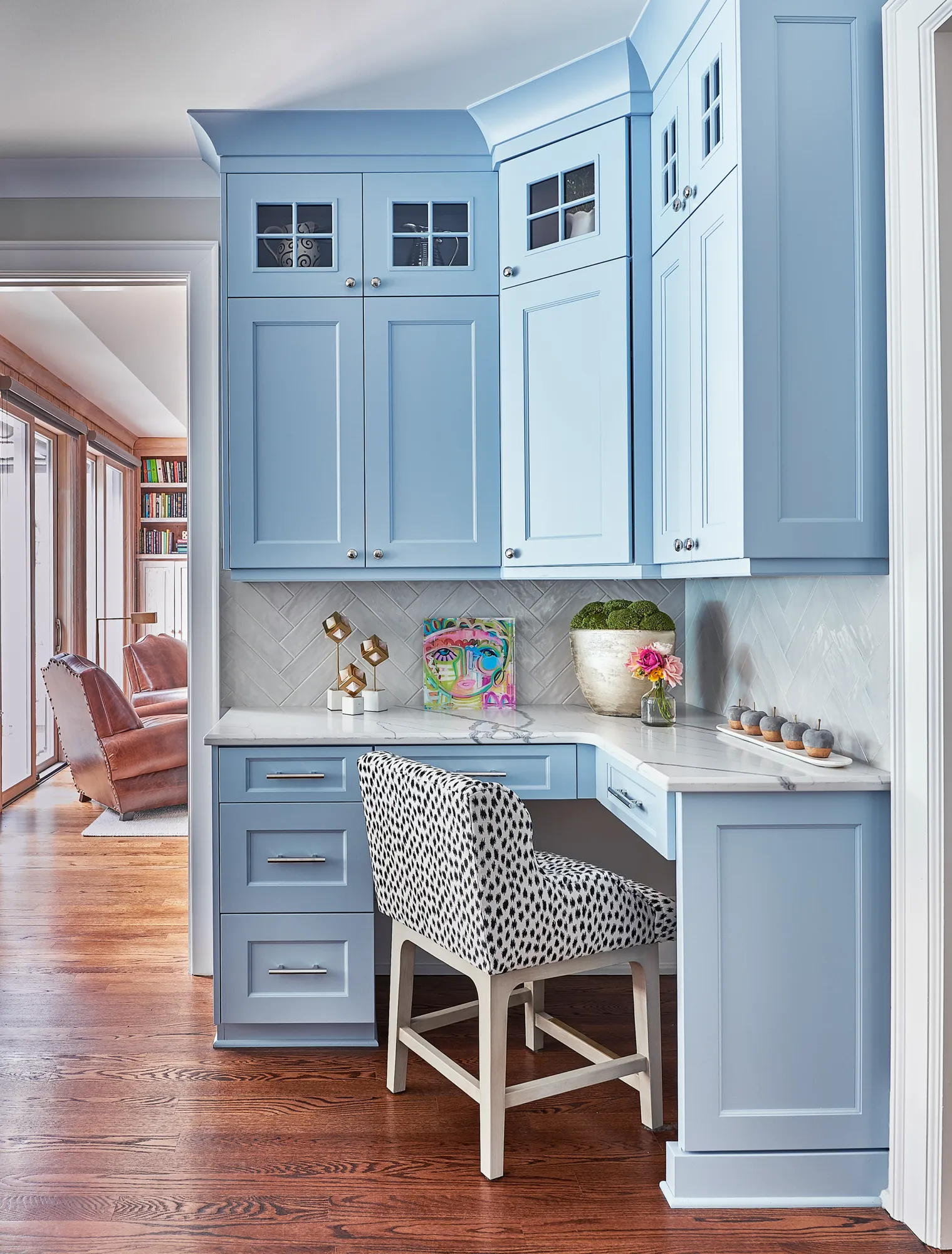 Light blue built-in cabinetry forms a home office nook with a white marble desk and a spotted chair. The space opens to a reading room with leather chairs.