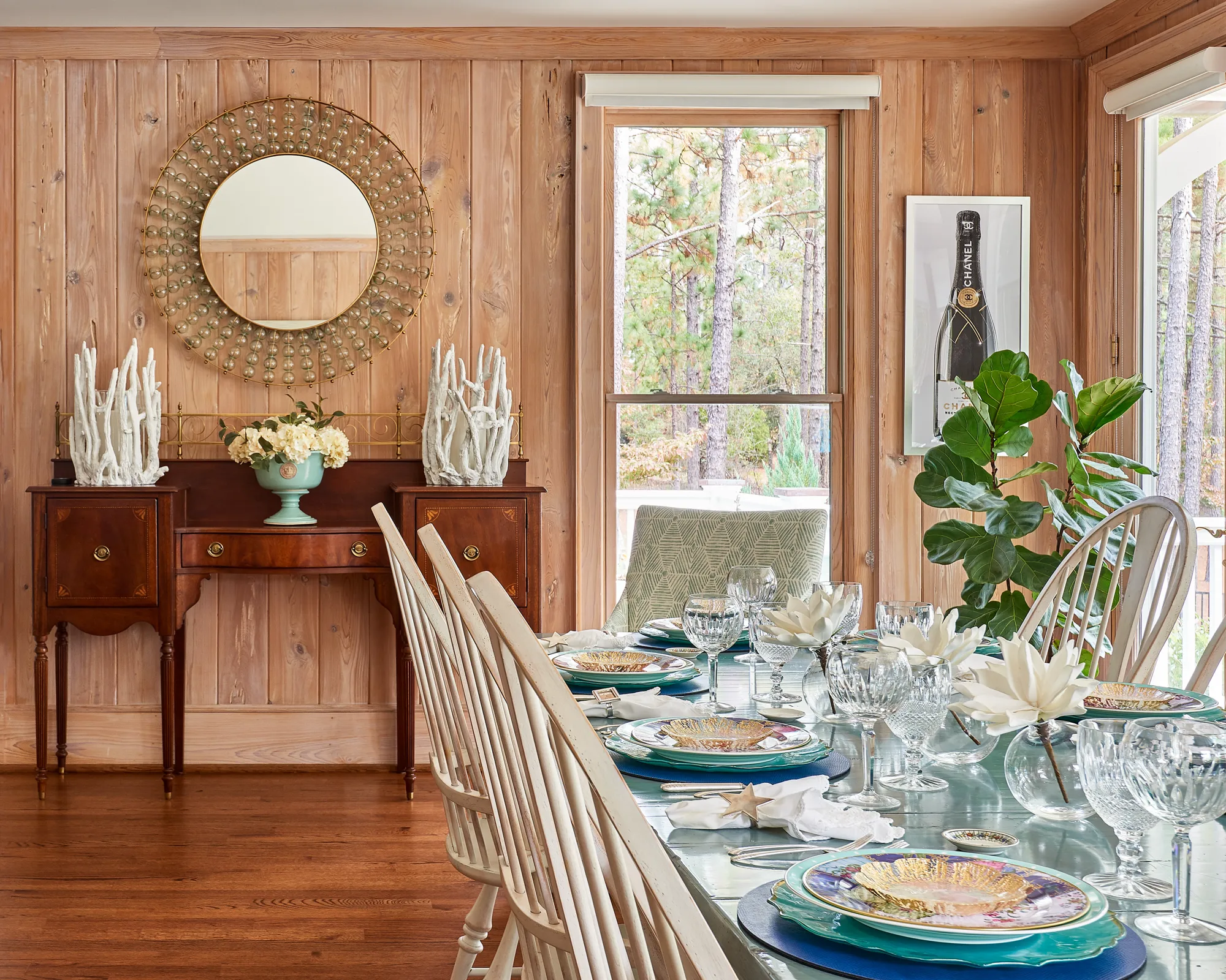 An elegant dining area with pine paneled walls and dark wood floors. The table is set with blue placemats, gold plates, crystal glasses, and white flowers.