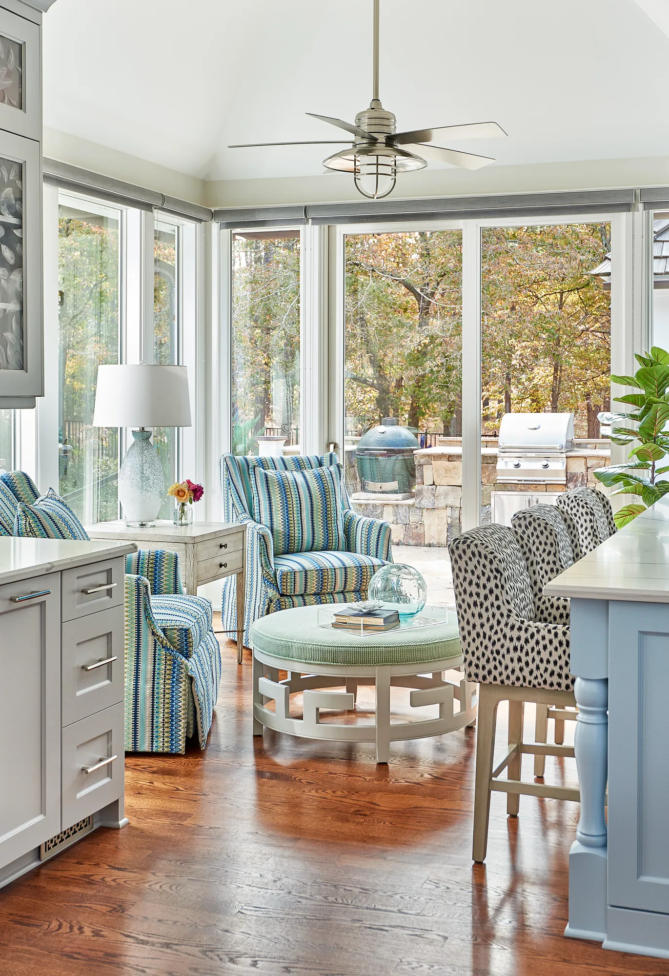 A cheerful interior space with blue cabinets and striped chairs looks out onto an outdoor kitchen and patio through large windows, integrating indoor and outdoor living.