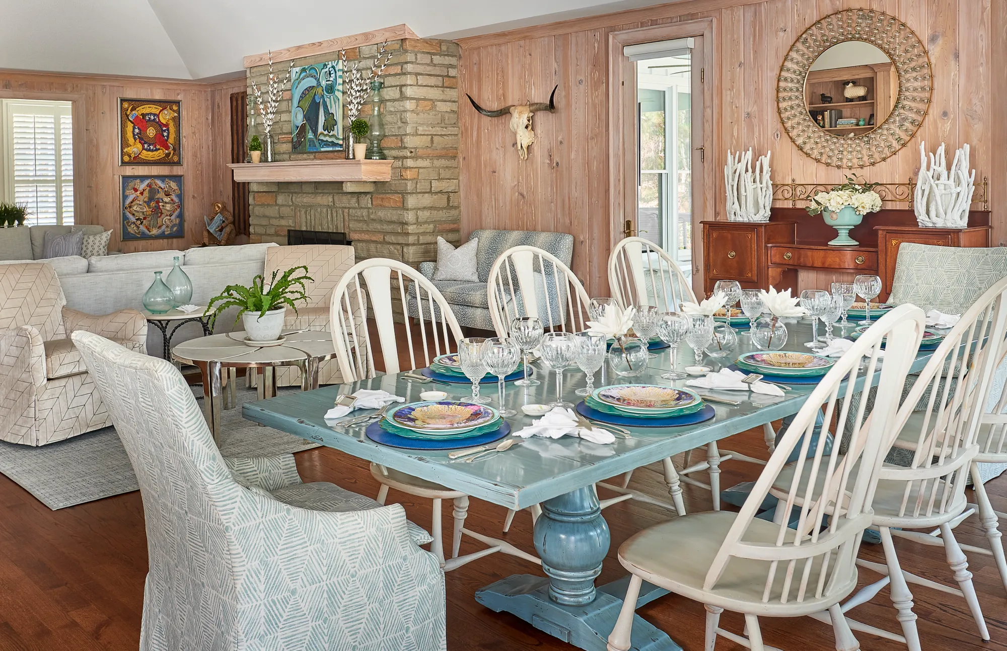 A spacious, open-concept dining area with wood panel walls and a stone fireplace. A distressed blue dining table is formally set with glassware and colorful plates, surrounded by mixed chairs.
