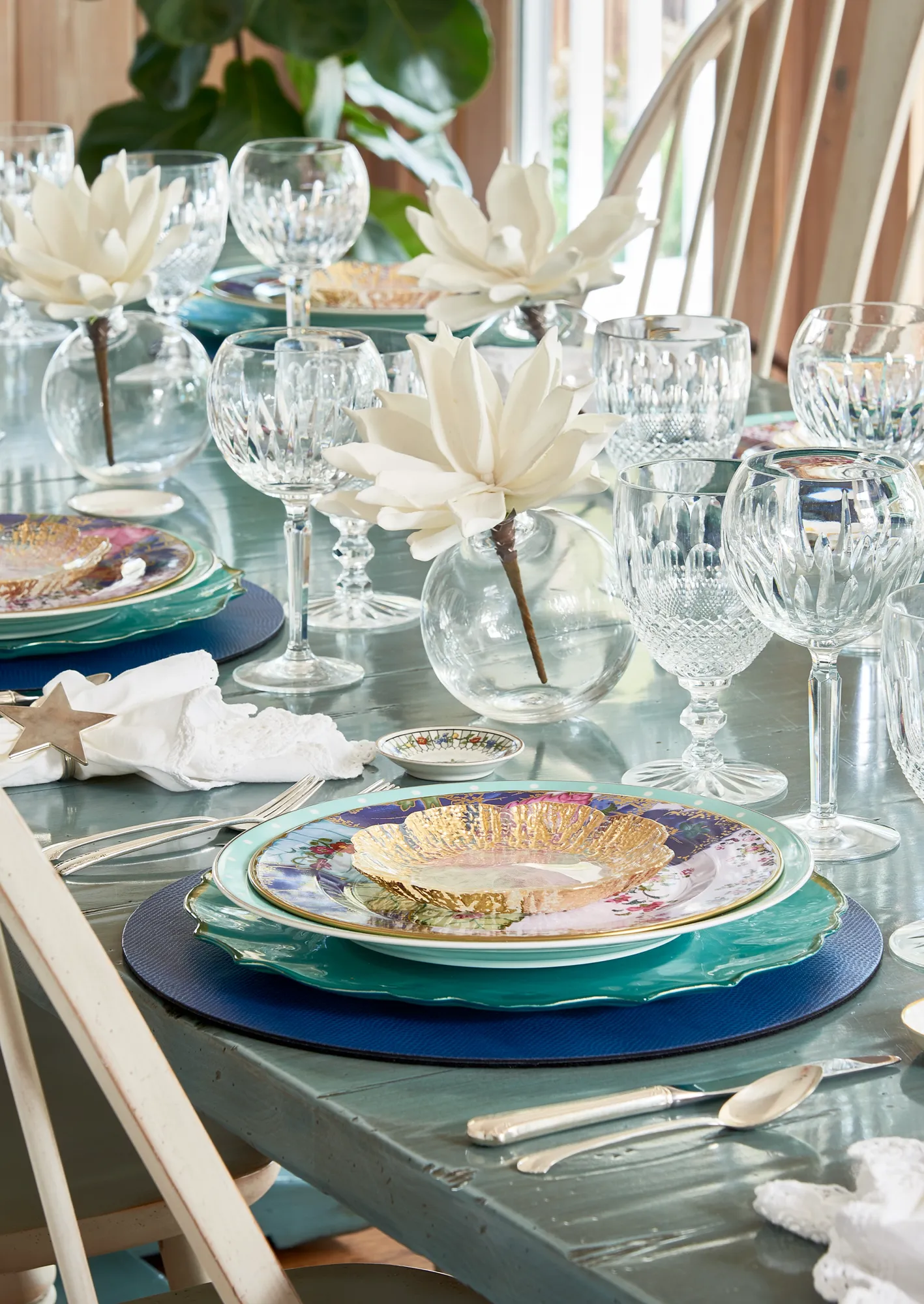 A formal dining table is decorated with layered blue and gold dishes, crystal stemware, silver utensils, and white decorative flowers on a teal painted wood table.