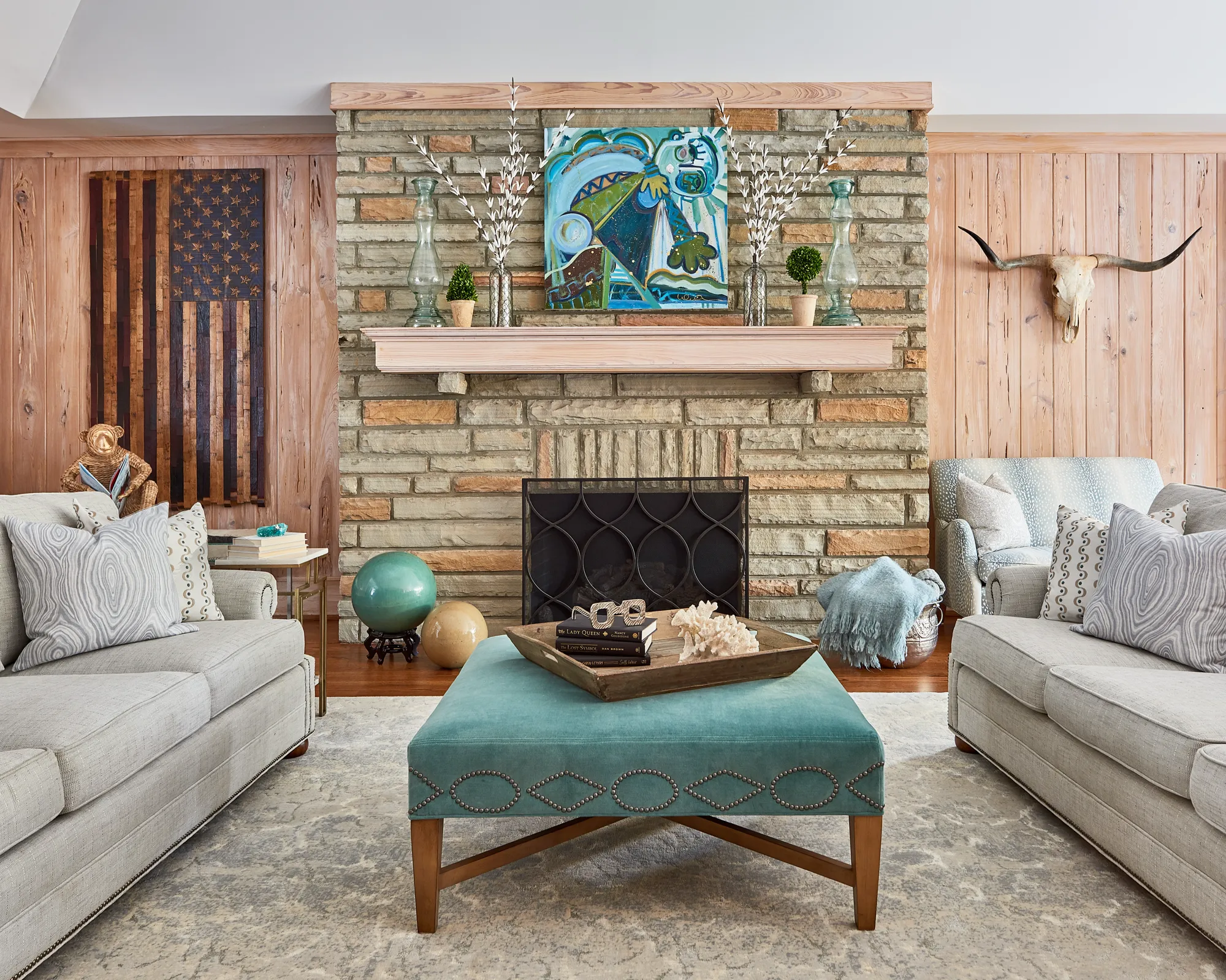 A rustic-chic living room with a stone fireplace and wood panel walls. Gray sofas face a studded turquoise velvet ottoman centered on a patterned rug.