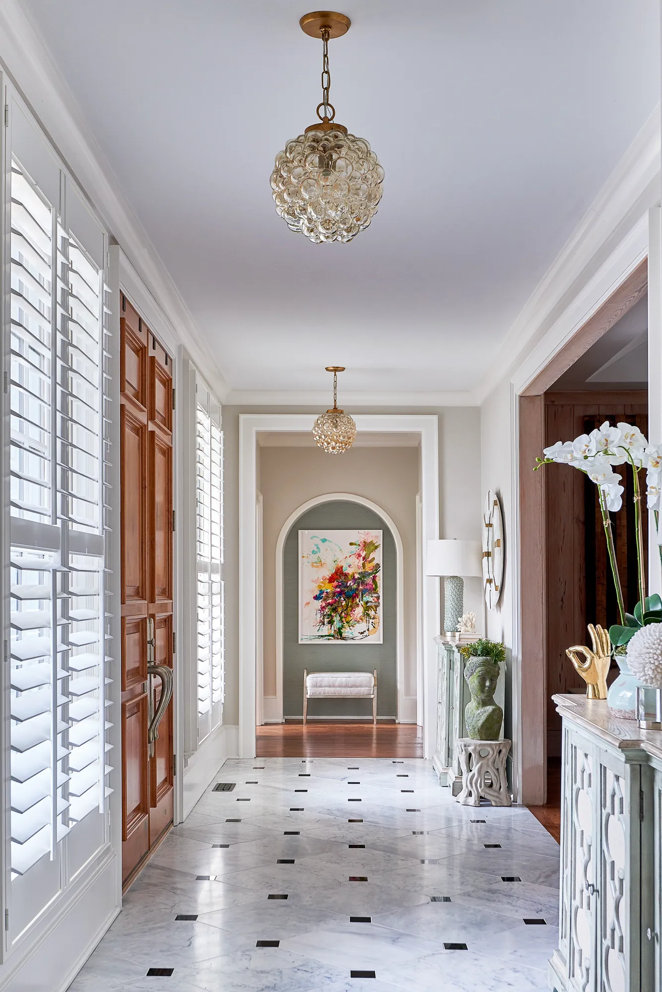 A classic residential hallway with a white marble floor and black inlays. A brown door and plantation shutters are on the left, leading past a decorated console and abstract painting.