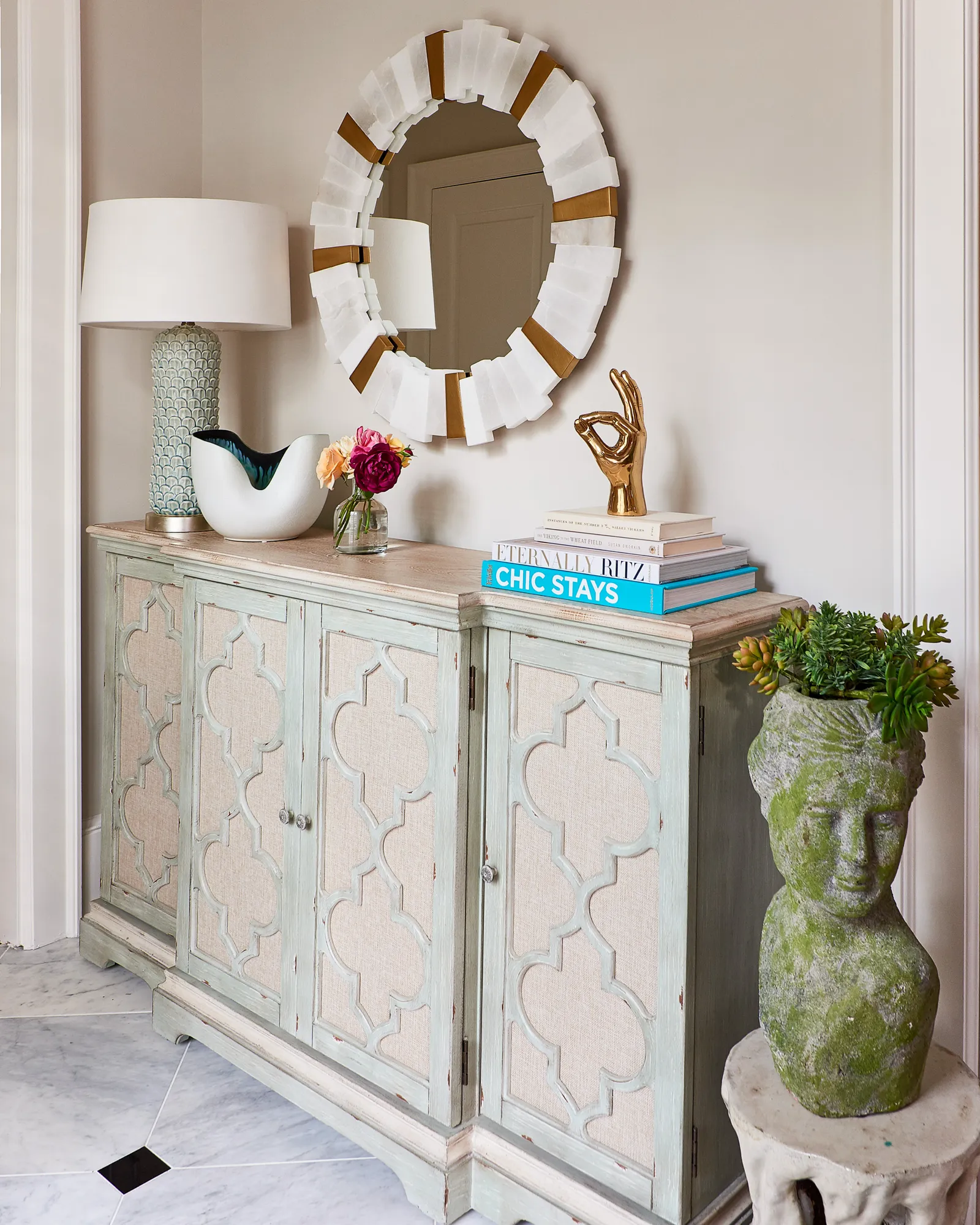 A stylish distressed blue credenza featuring quatrefoil fabric panels, topped with a white lamp, flowers, and stacked books. A gold and white sunburst mirror hangs above.