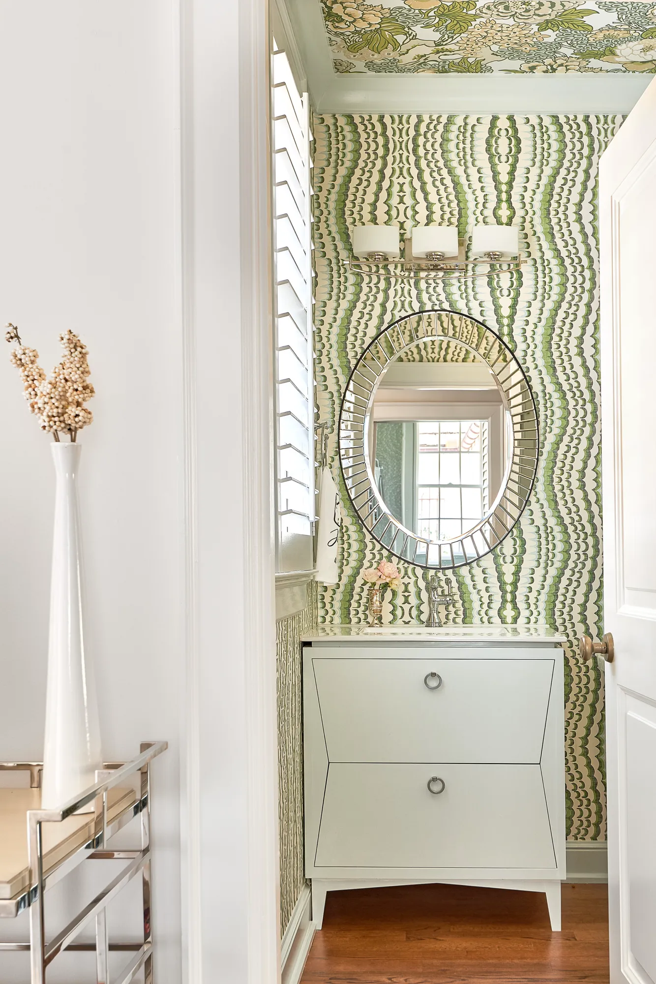 A view into a stylish powder room featuring green scalloped wallpaper, a contrasting floral ceiling, a white modern vanity, and a large oval segmented mirror.
